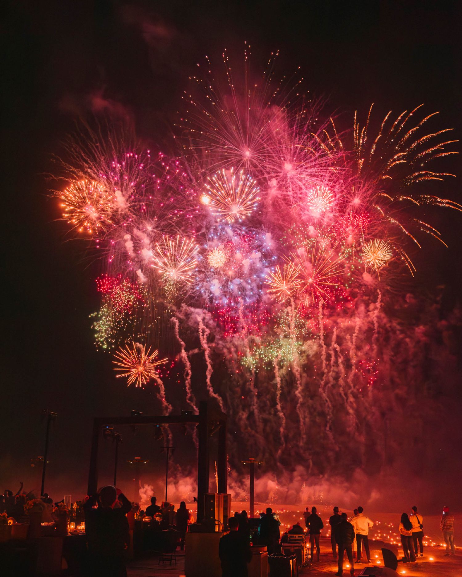 Colorful fireworks display lighting up the night sky in red and pink hues as silhouetted spectators watch from below.