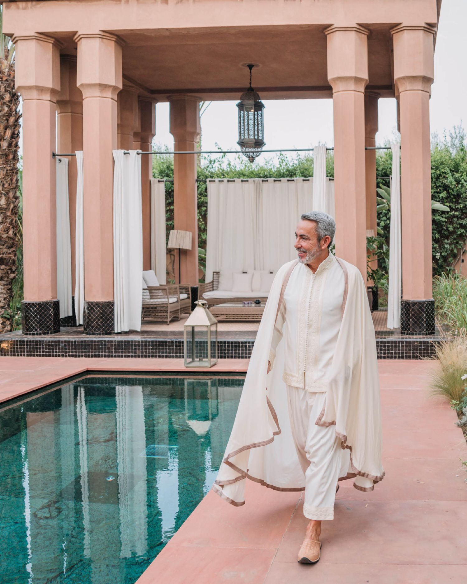 Person in white attire walks along pink poolside terrace with elegant columns and hanging lantern at luxury resort.