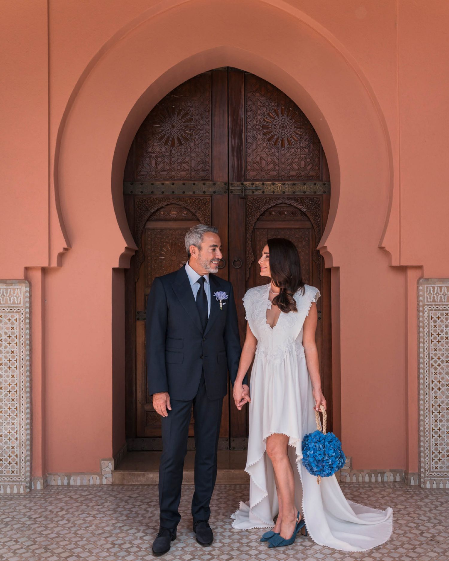 A couple stands in front of an ornate Moroccan archway with decorative wooden doors and terracotta walls.