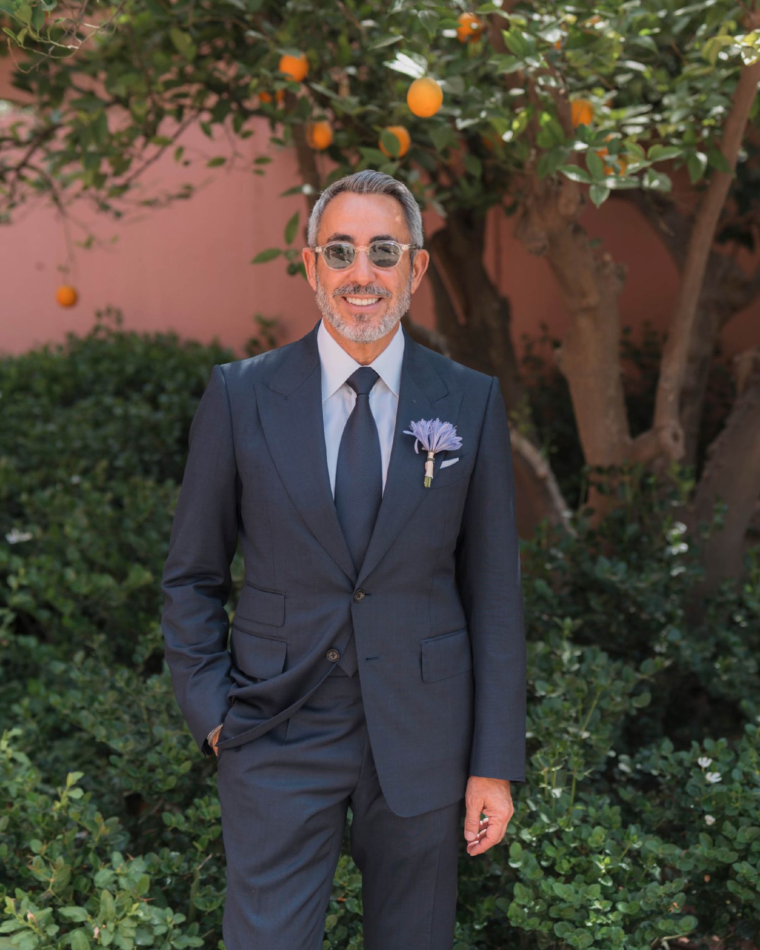 Someone in a navy blue suit with sunglasses stands smiling in front of an orange tree and pink wall.