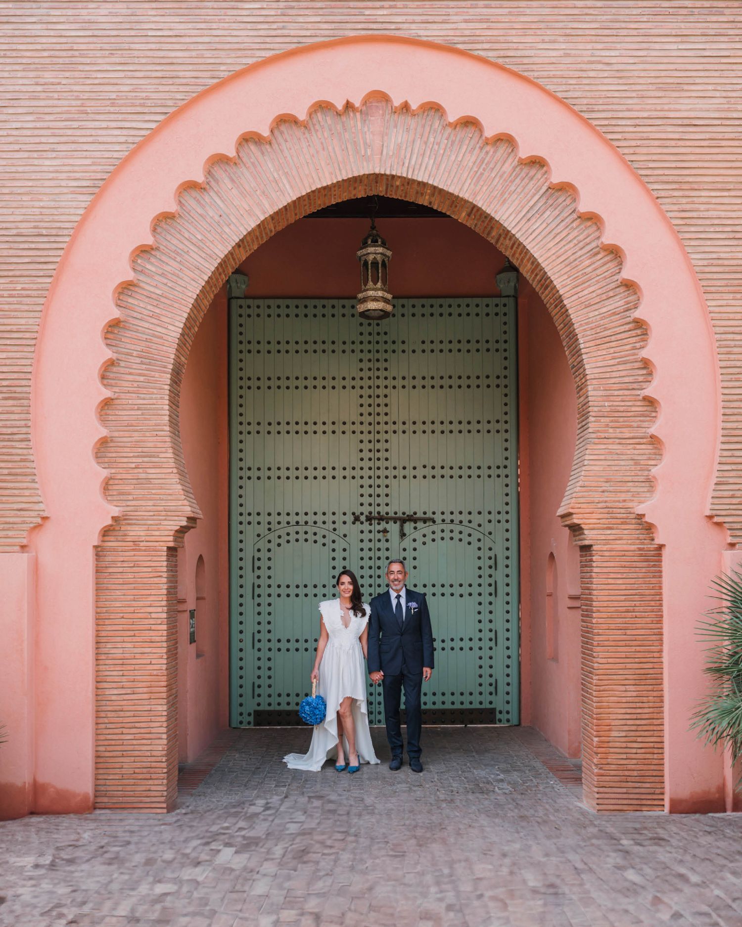 A couple poses beneath an ornate pink Moroccan archway with traditional studded green doors.