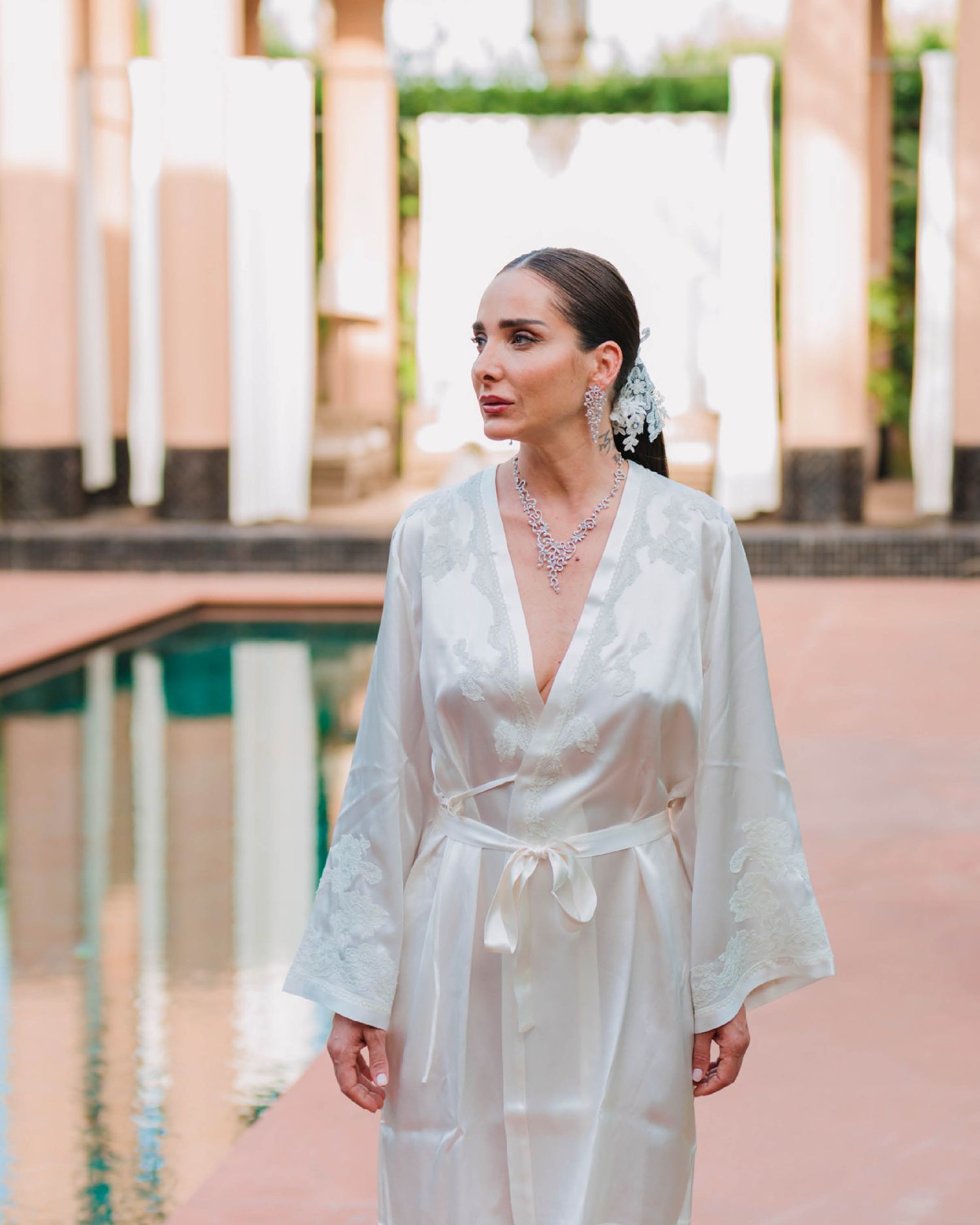 A person wearing an elegant white silk robe with long sleeves stands near a swimming pool in a luxurious setting.