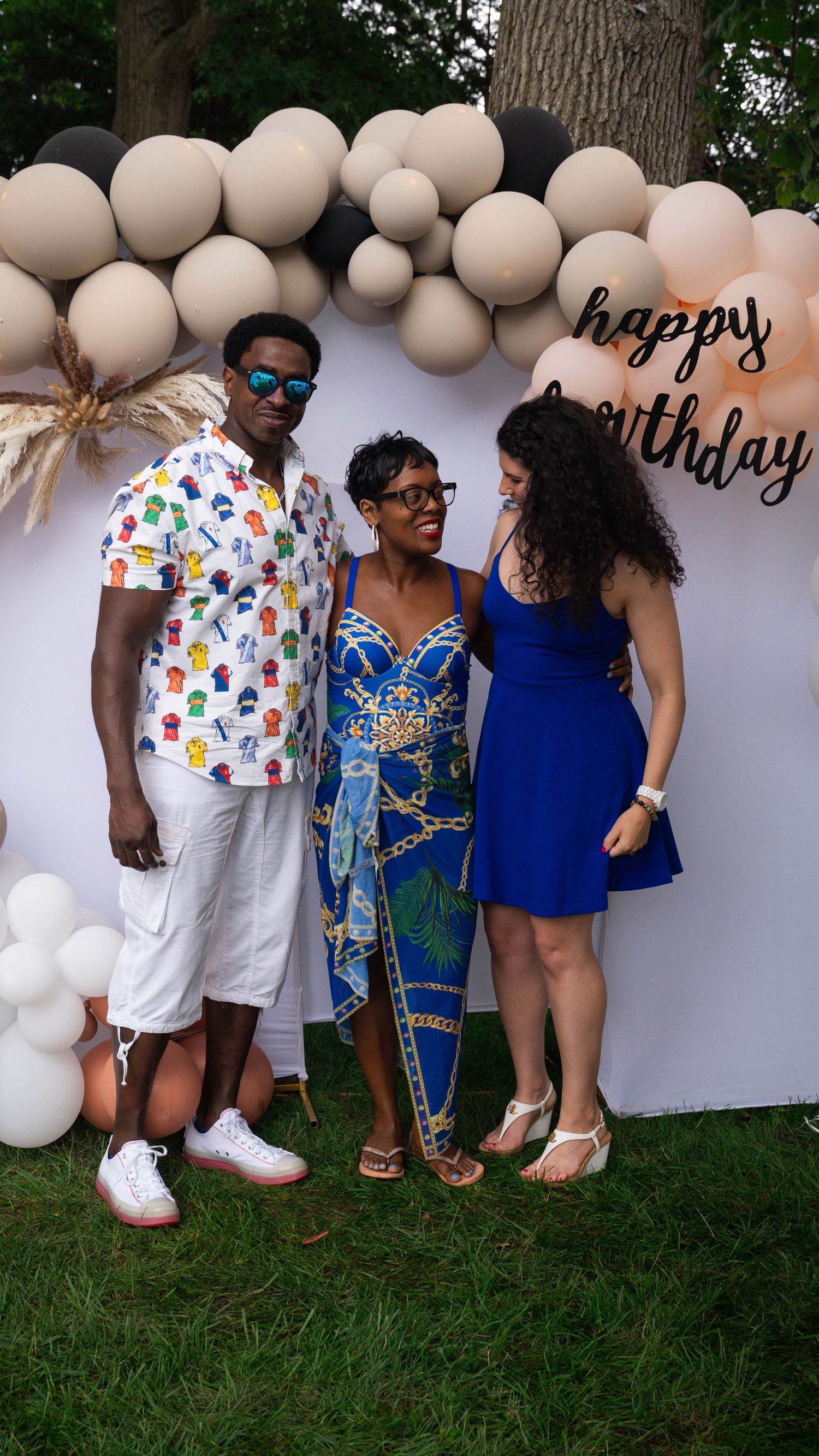 Birthday celebration on lawn with balloon arch decor and people in colorful summer outfits gathered for a photo.