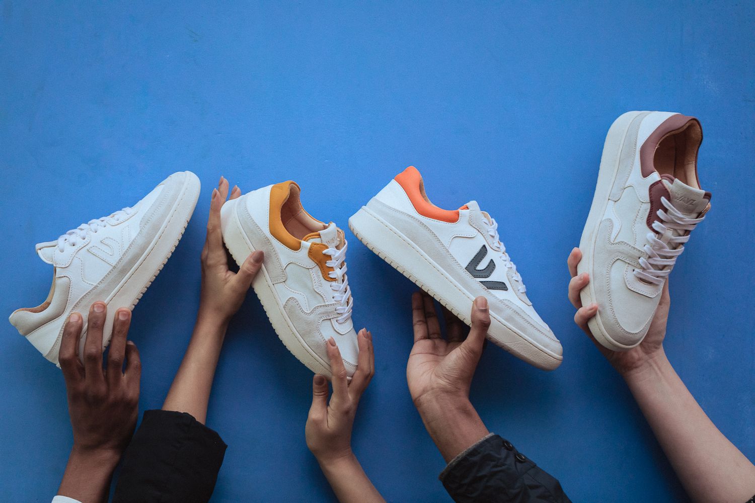Multiple hands holding up white athletic sneakers with orange and brown accents against a blue background.