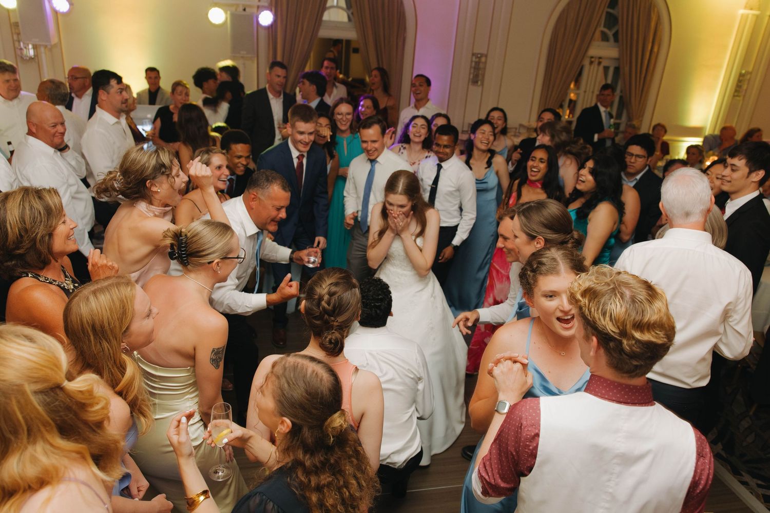 Wedding guests surround a couple dancing at a reception in an elegant ballroom with warm lighting.