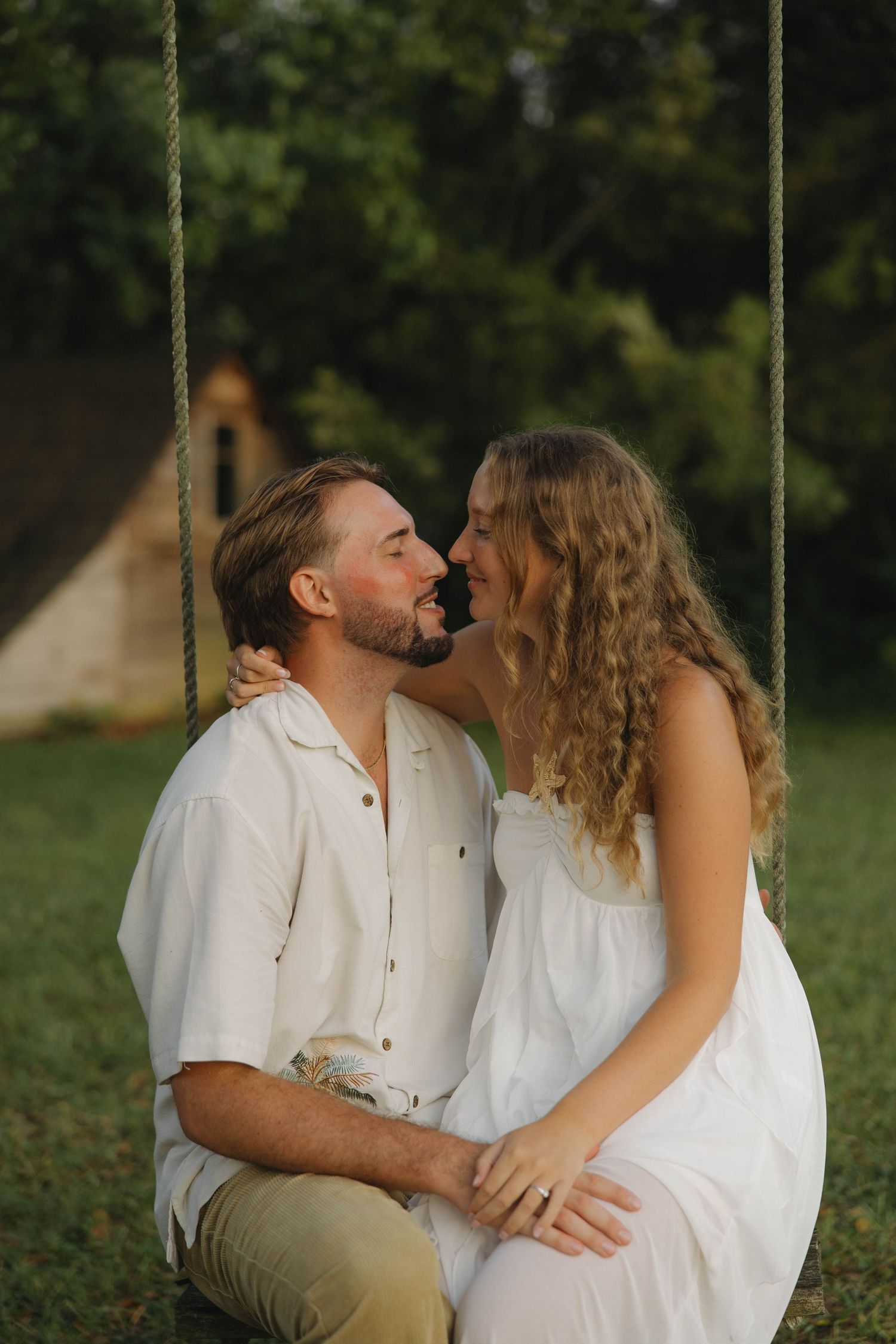 A romantic couple in white outfits share intimate moments while sitting on a swing outdoors.