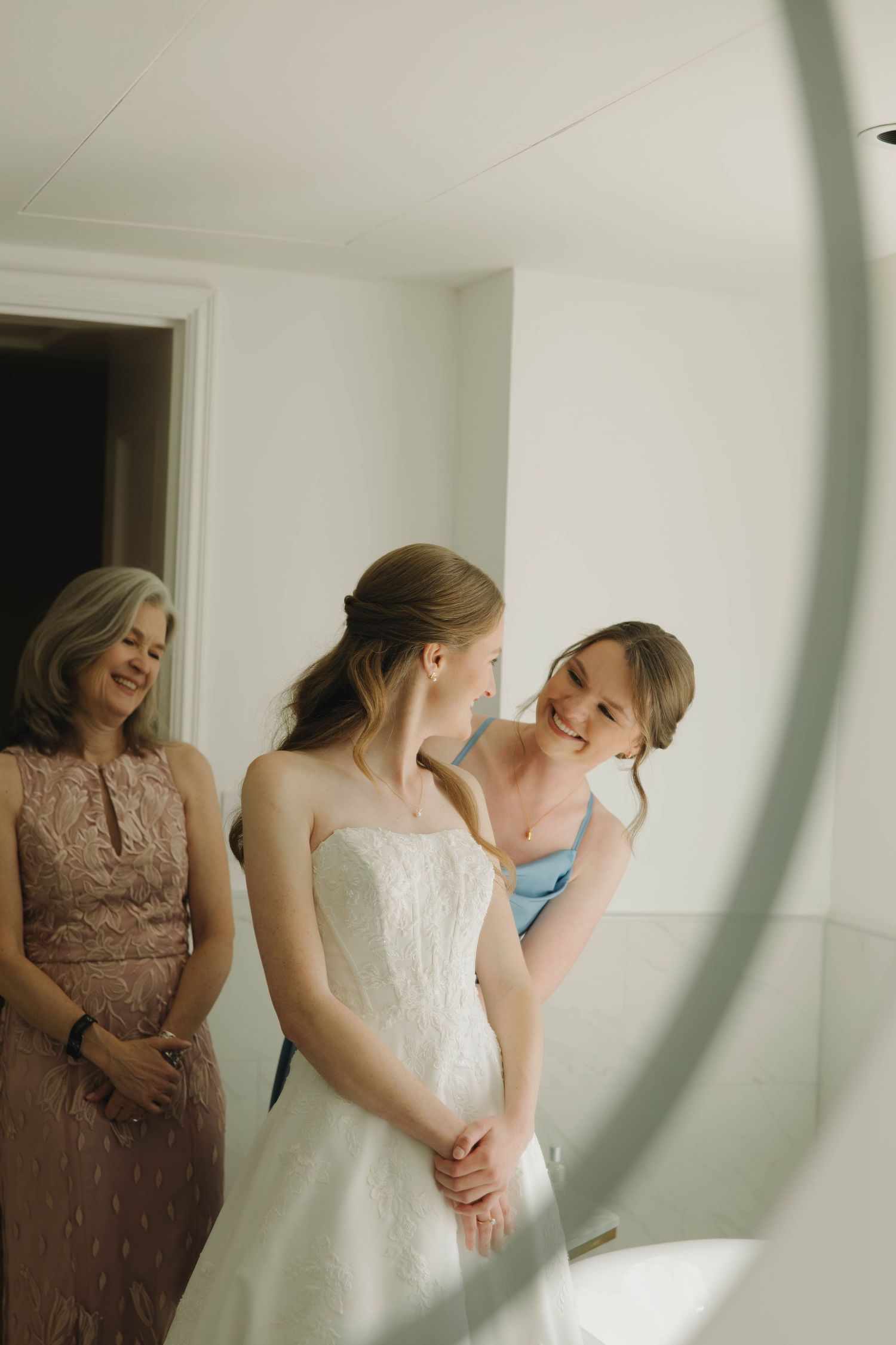 A candid moment shows people laughing and embracing in a bright white room during wedding preparations.