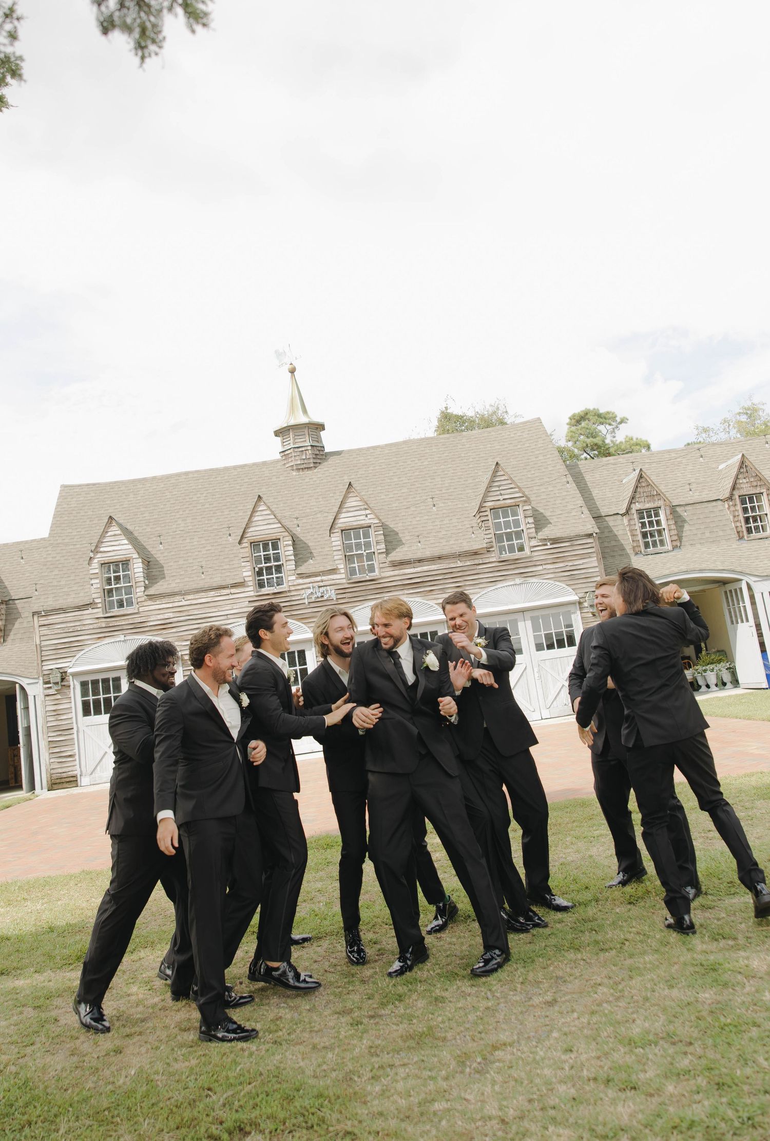 Groomsmen in black suits playfully pose and jump together on grass in front of a classic stone building with dormers.