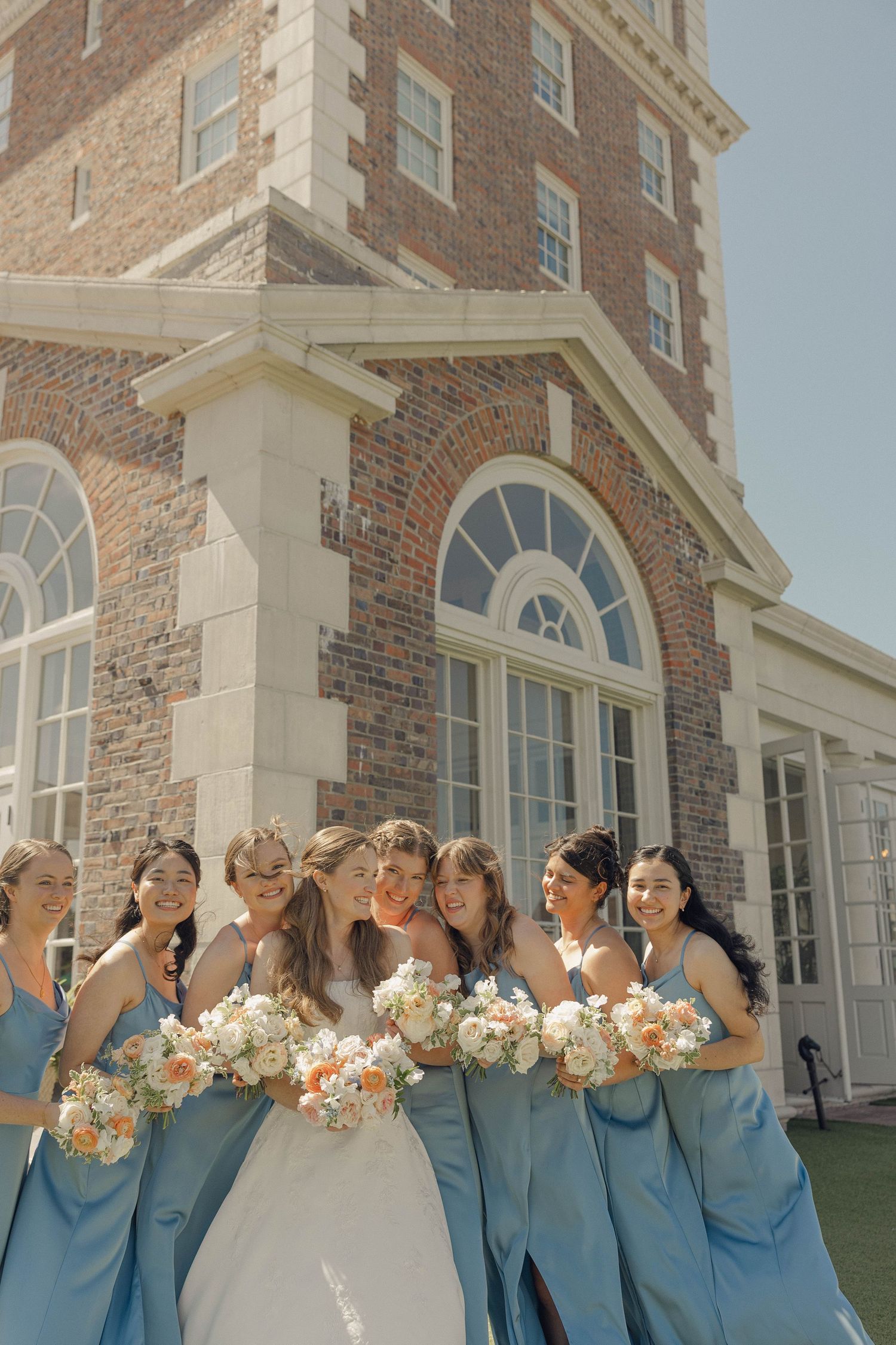 A wedding party in blue dresses poses together while holding peach and white floral bouquets outside a brick building.