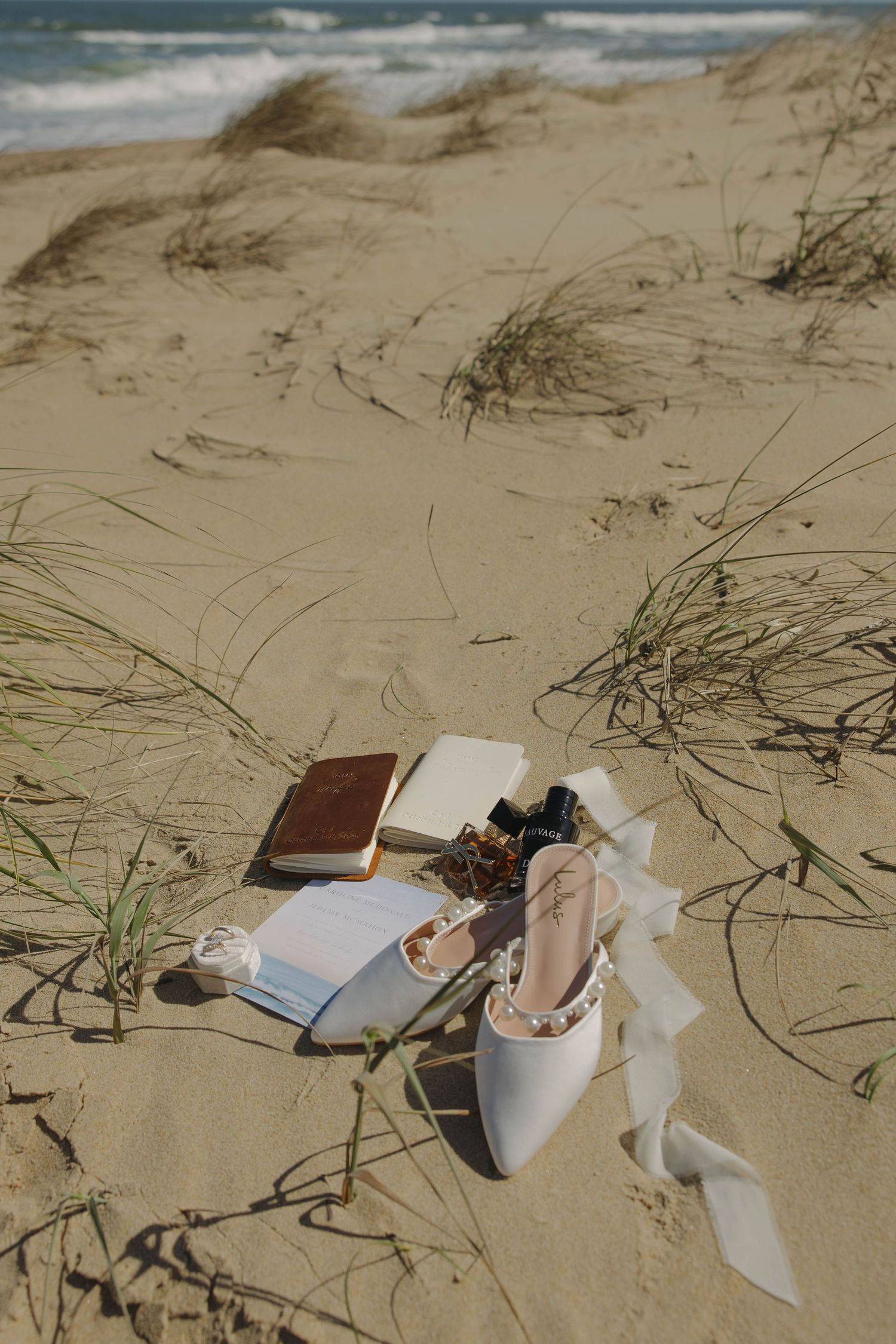 Wedding rings and ribbons rest on sandy beach dunes with scattered beach grass and ocean waves in background.