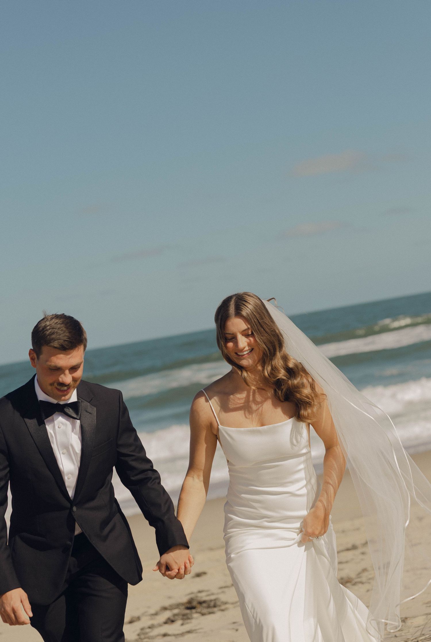 A joyful couple in wedding attire walks hand in hand along a scenic beach with waves crashing in the background.