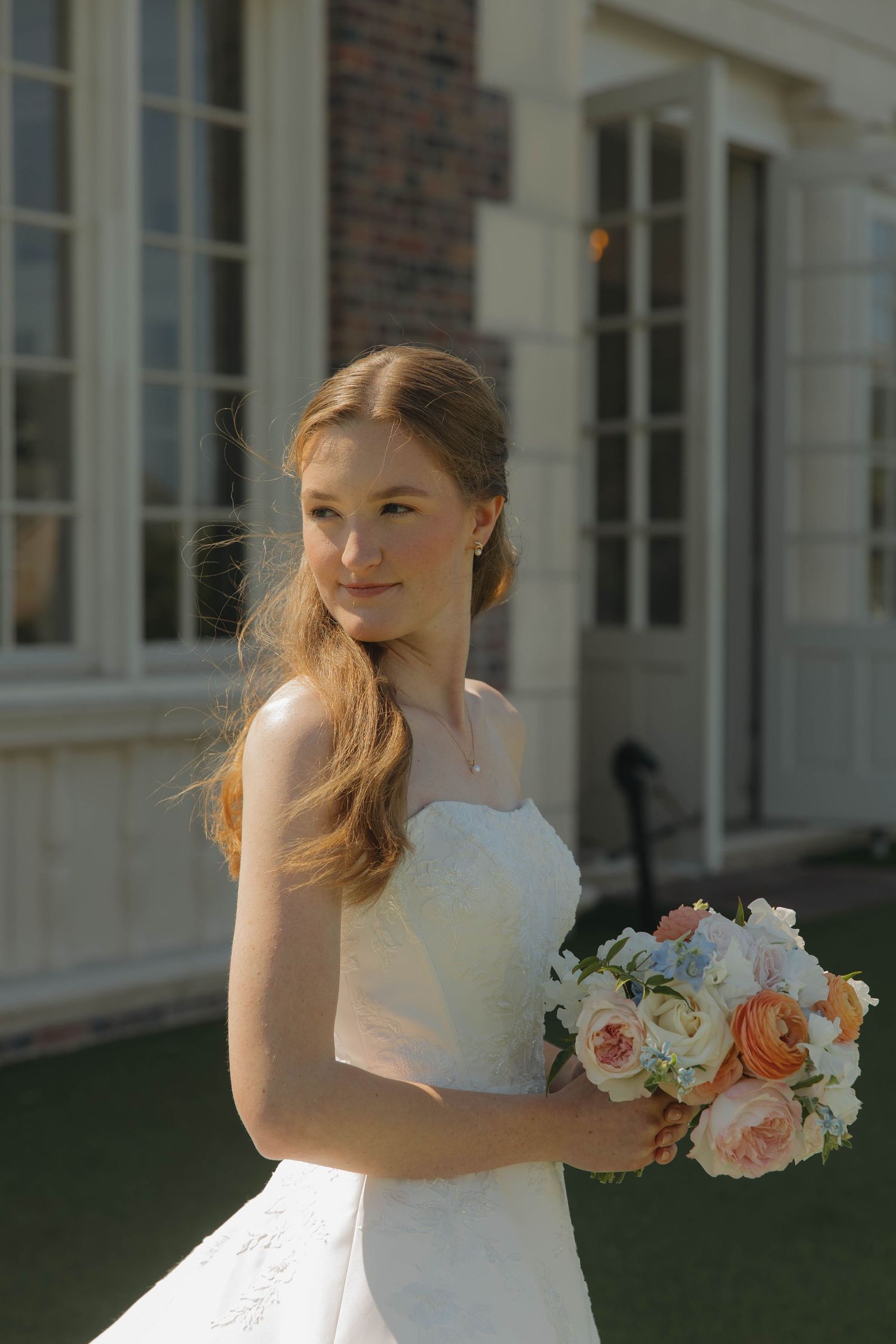 A bride in a white strapless wedding dress holds a peach and white rose bouquet outside a brick building.