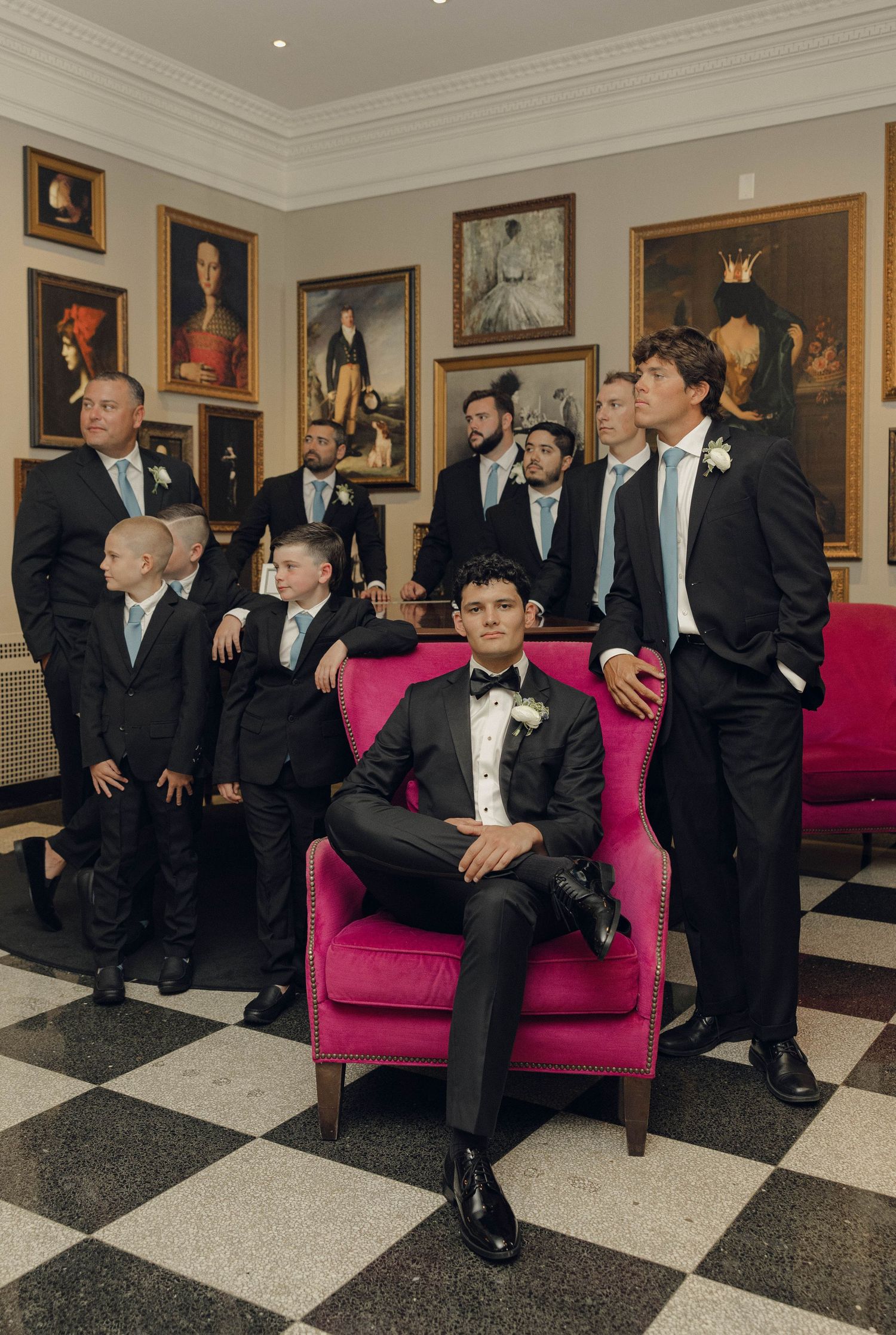 Groomsmen in black tuxedos gather and relax on a pink sofa in an elegant hallway with framed artwork on the walls.