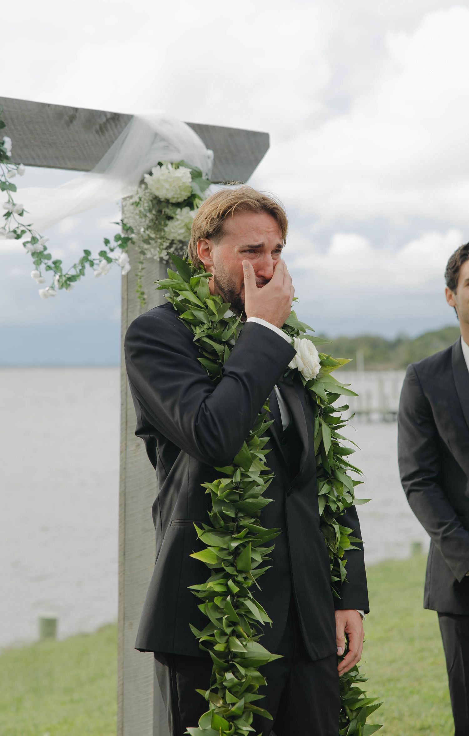 Groomsmen in black suits and ties stand in a row outdoors wearing white floral boutonnieres.