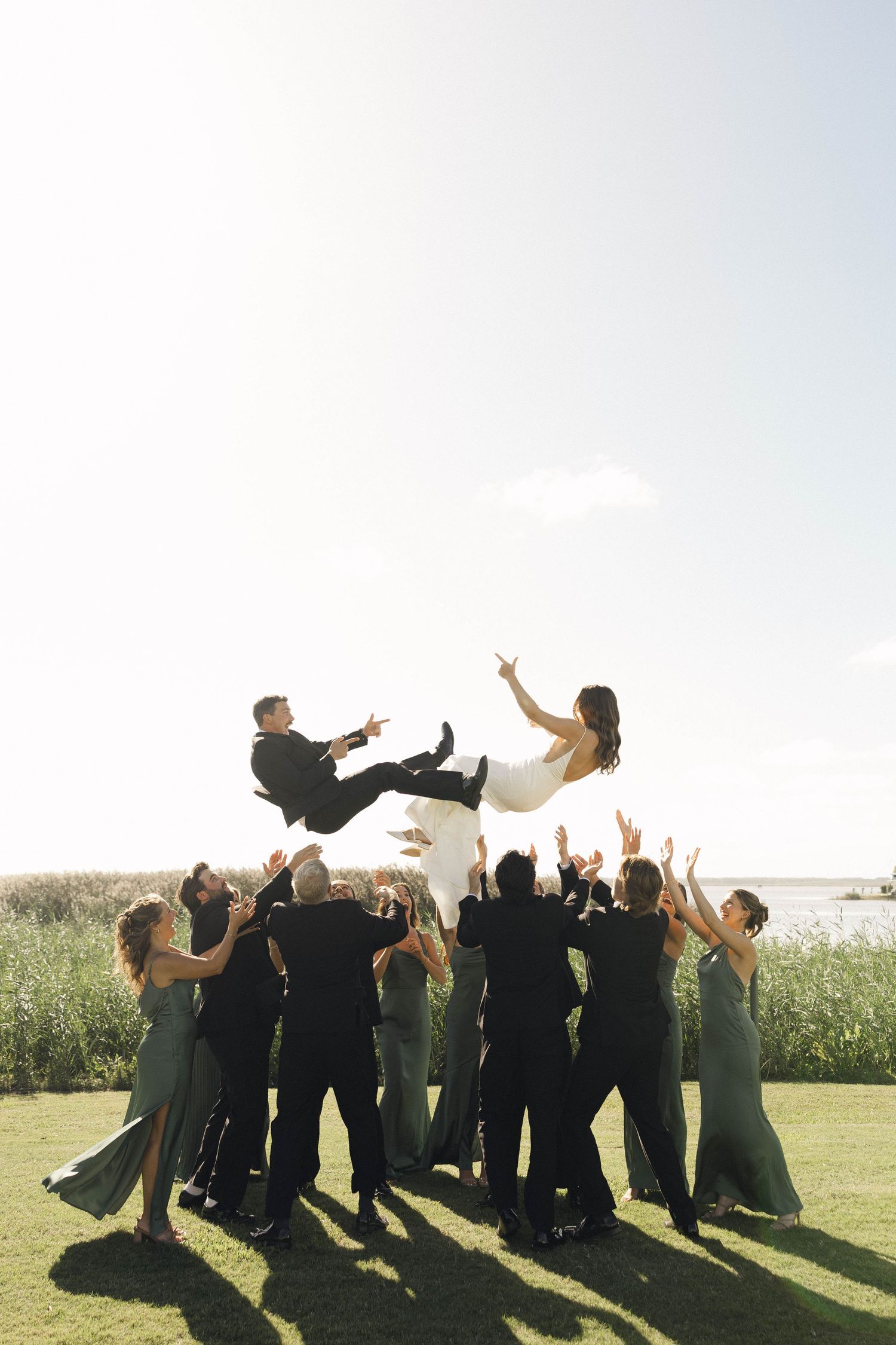 Wedding party jumps joyfully in unison on a sunny day in an outdoor field setting.