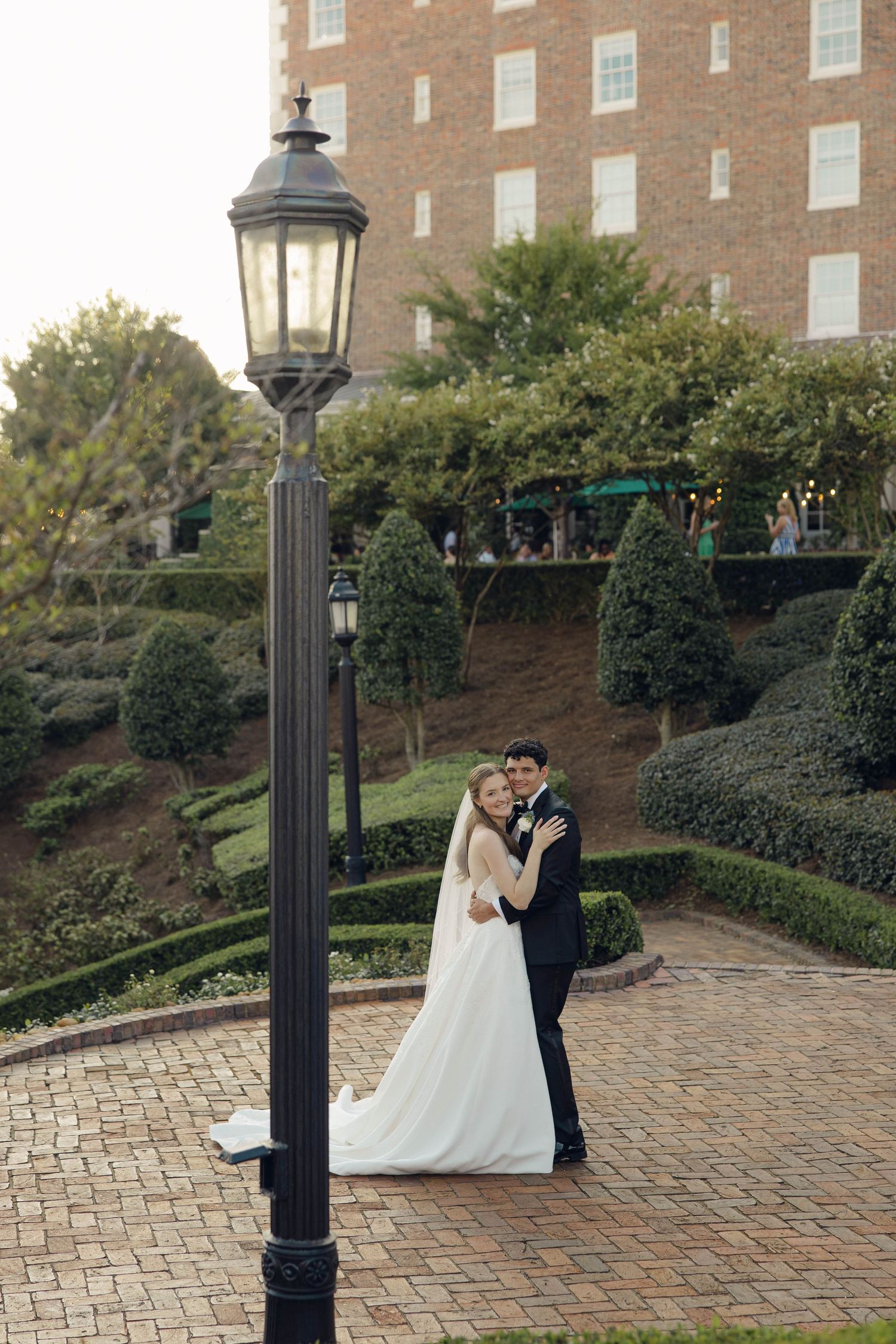 A couple shares a romantic moment on a brick pathway beside an ornate lamp post and manicured garden.