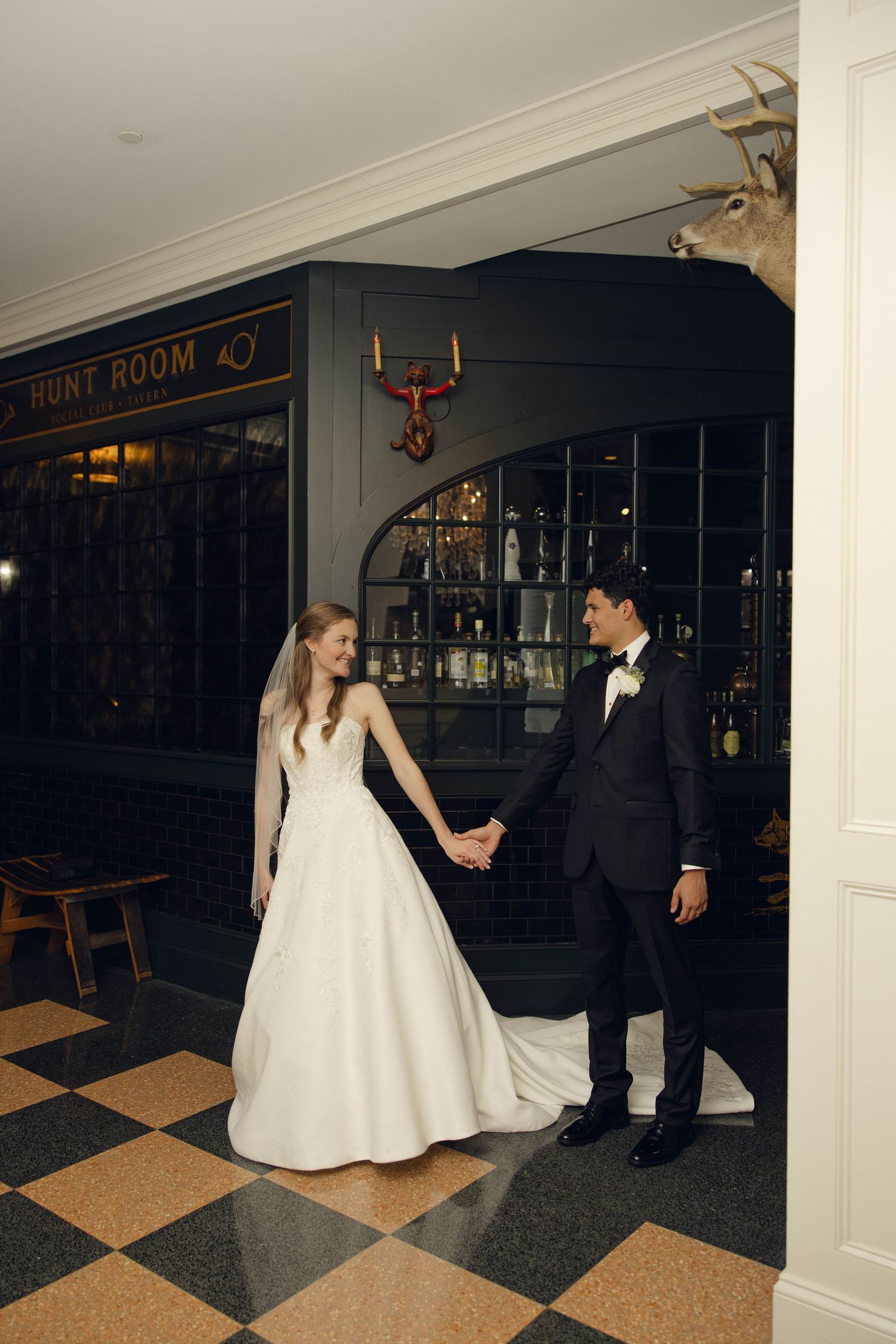 A couple in wedding attire stands in front of The Hunt Room entrance with checkerboard floors and rustic decor.
