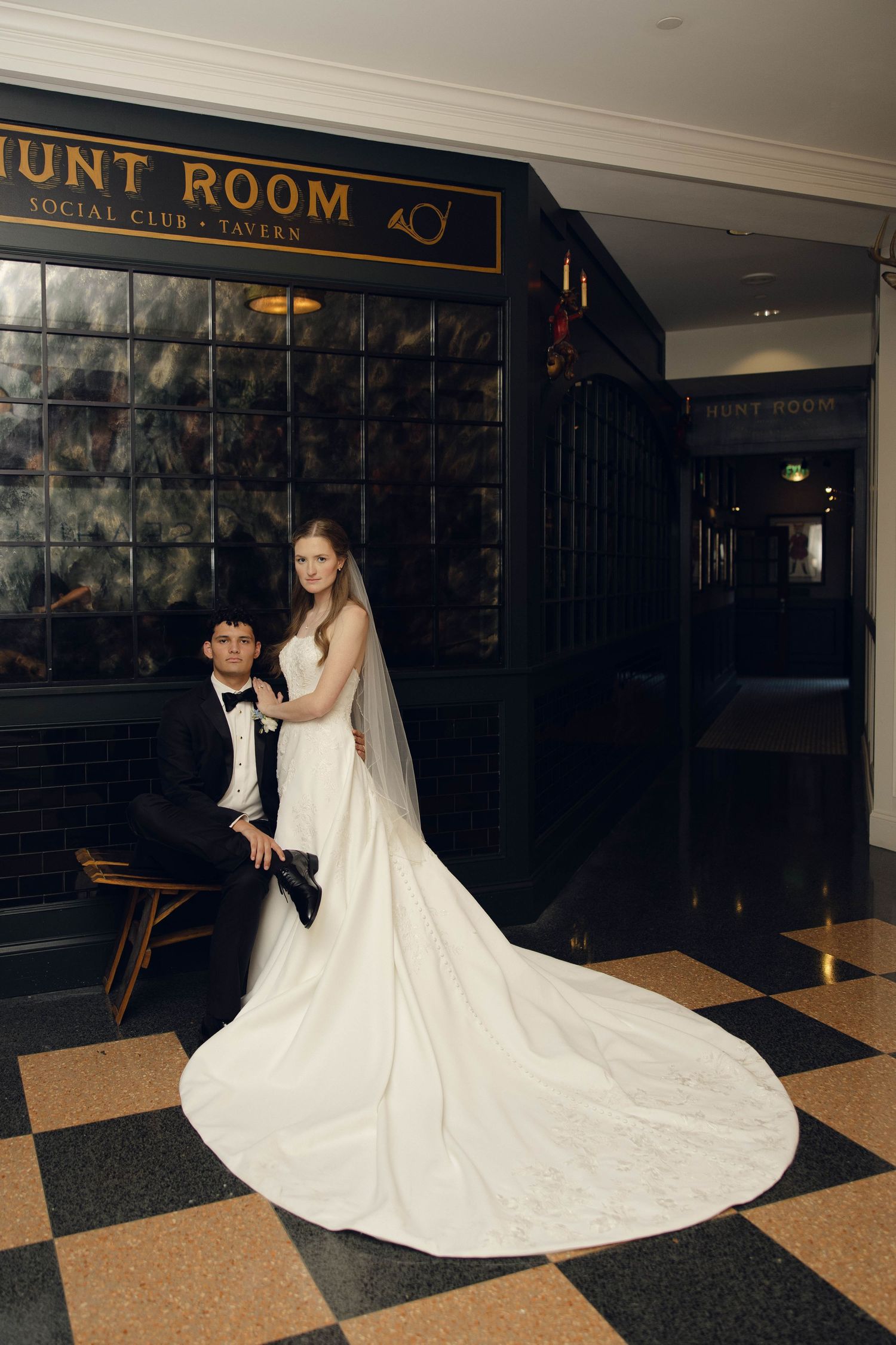 A couple in wedding attire poses in a dark hallway with a dramatic flowing white dress spread across the floor.