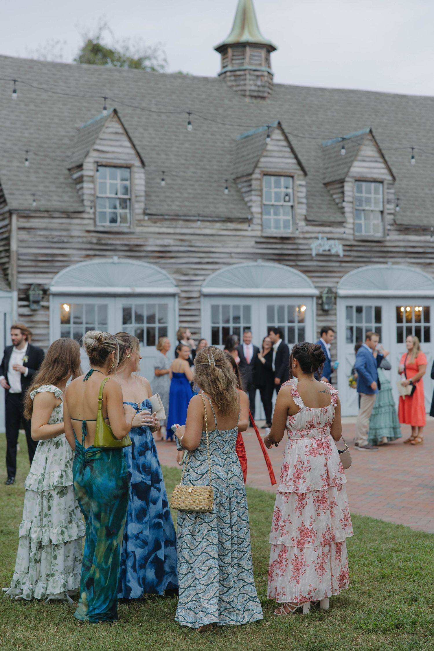 Group of wedding guests in colorful floral dresses gathered outside a charming venue with dormers and grey shingles.