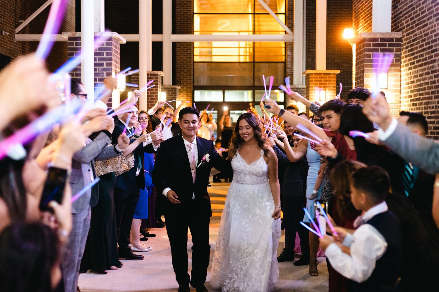 Wedding couple exits venue through glowing sparkler tunnel at night while guests line walkway with illuminated wands.