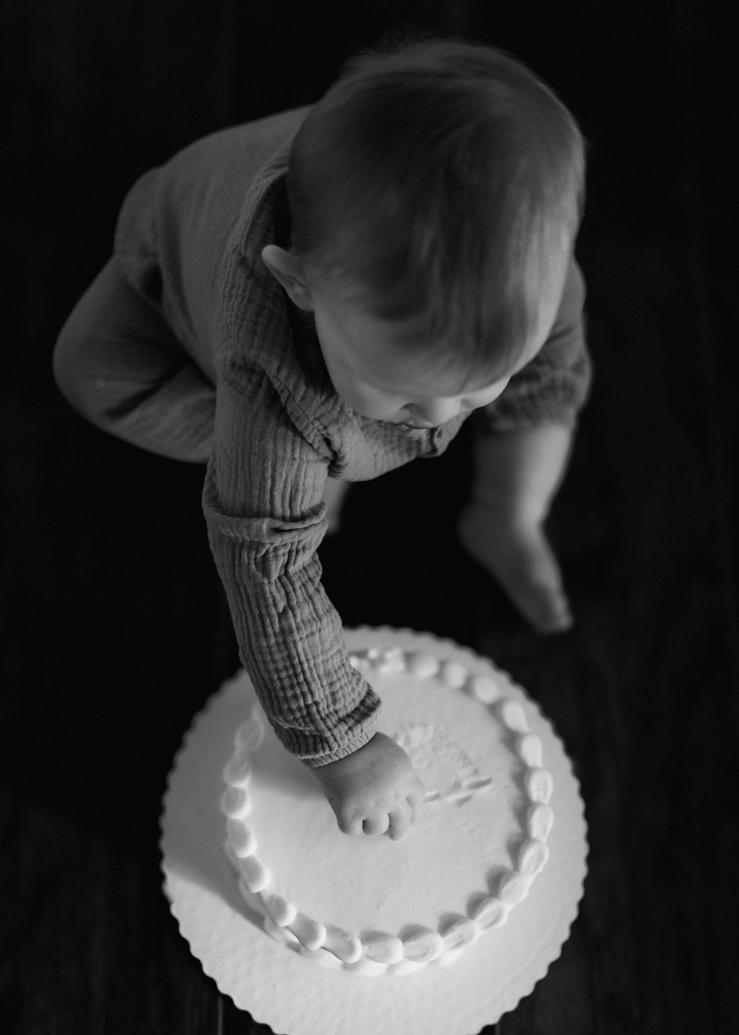 Black and white photograph of a small child bending down to touch a white frosted cake on a dark background.