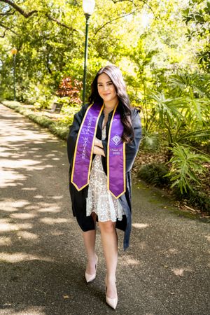Graduate in black gown and purple sash poses on garden pathway wearing heels.