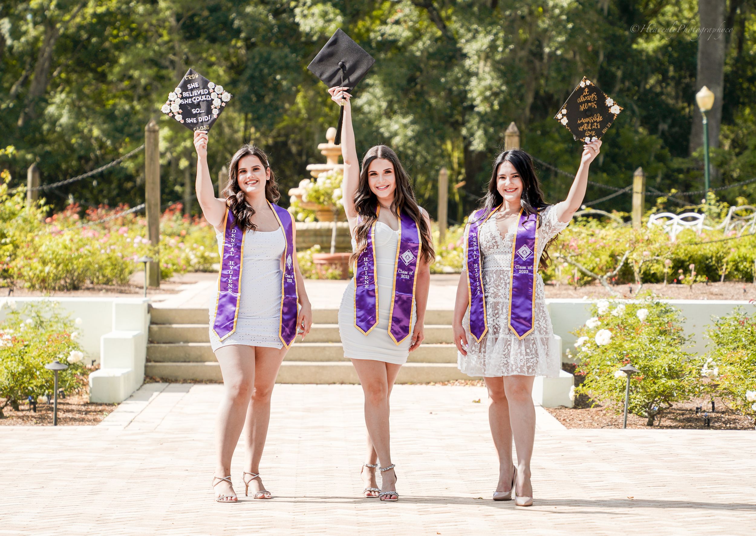 Graduates in white dresses and purple sashes pose together outdoors in a sunny garden setting.