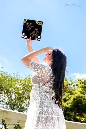 Graduate holds up graduation cap against bright blue sky in celebratory pose.