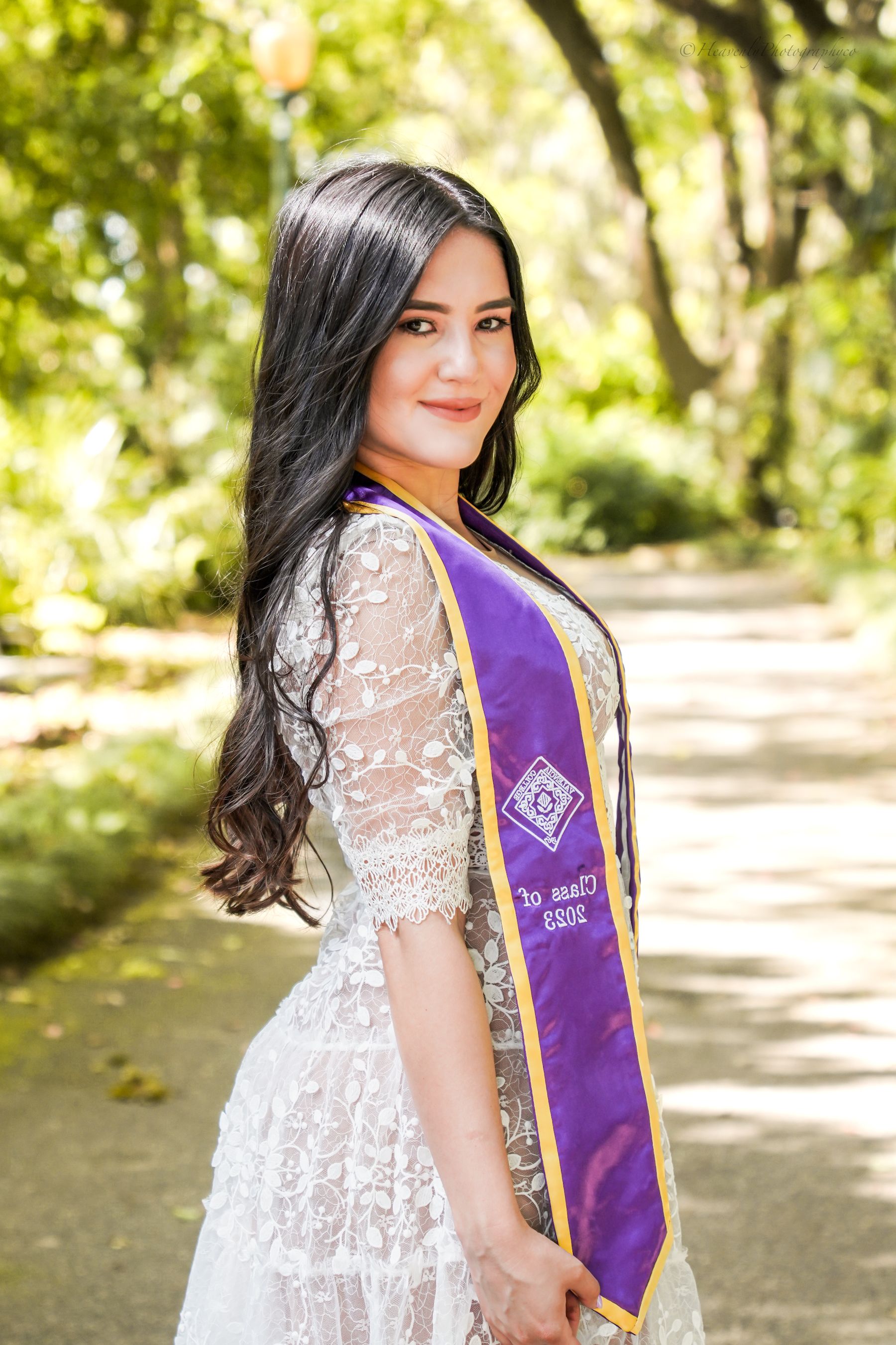 Graduate in white lace dress and purple sash poses in a sunlit garden pathway.