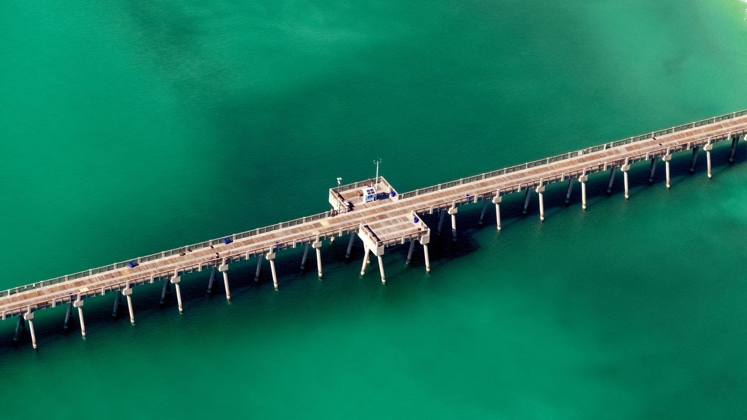 Aerial view of an industrial pier stretching across vibrant turquoise waters with a processing facility in the center.