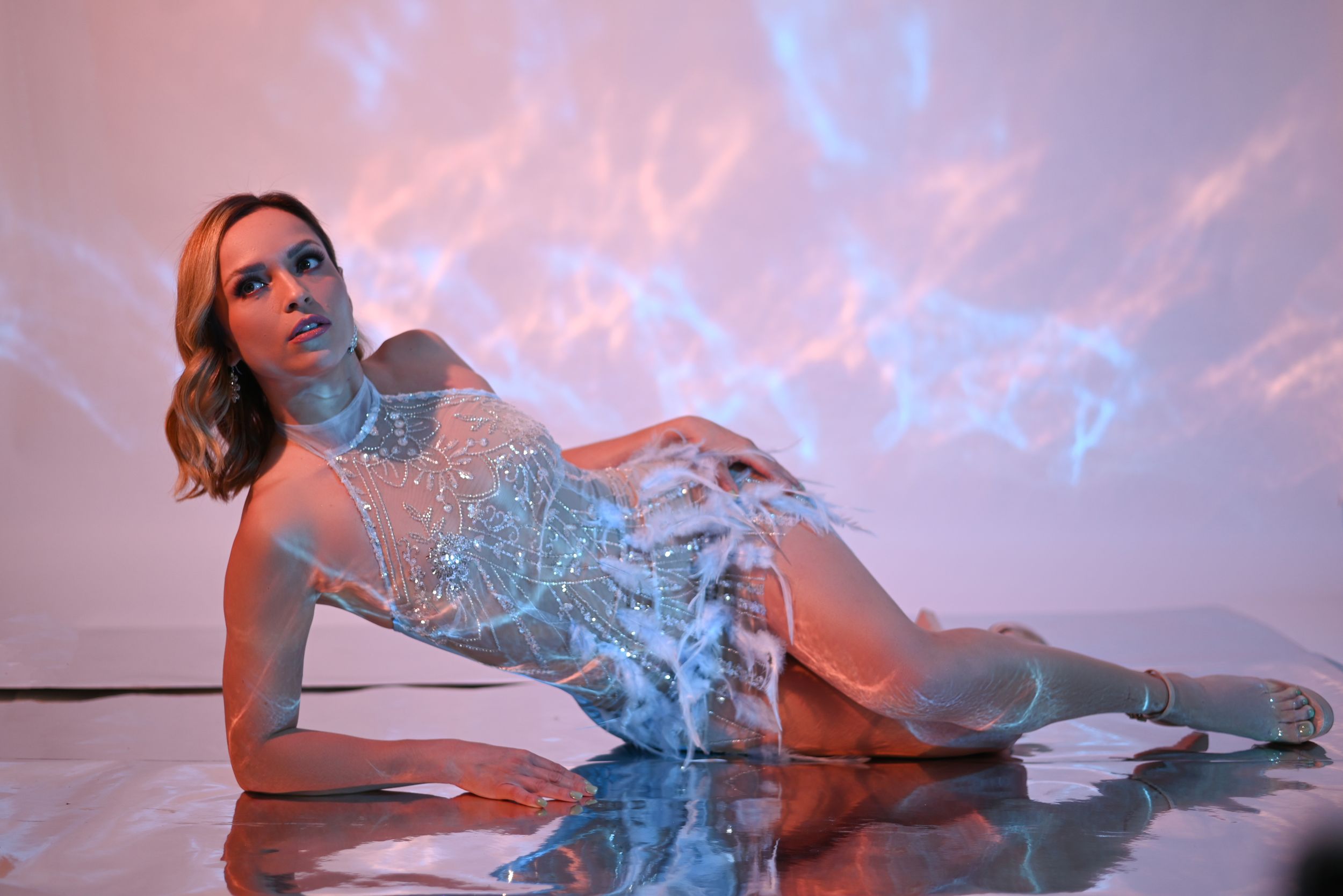Ethereal fashion photo with model in shimmering blue dress lying on reflective floor under pink and blue sky.