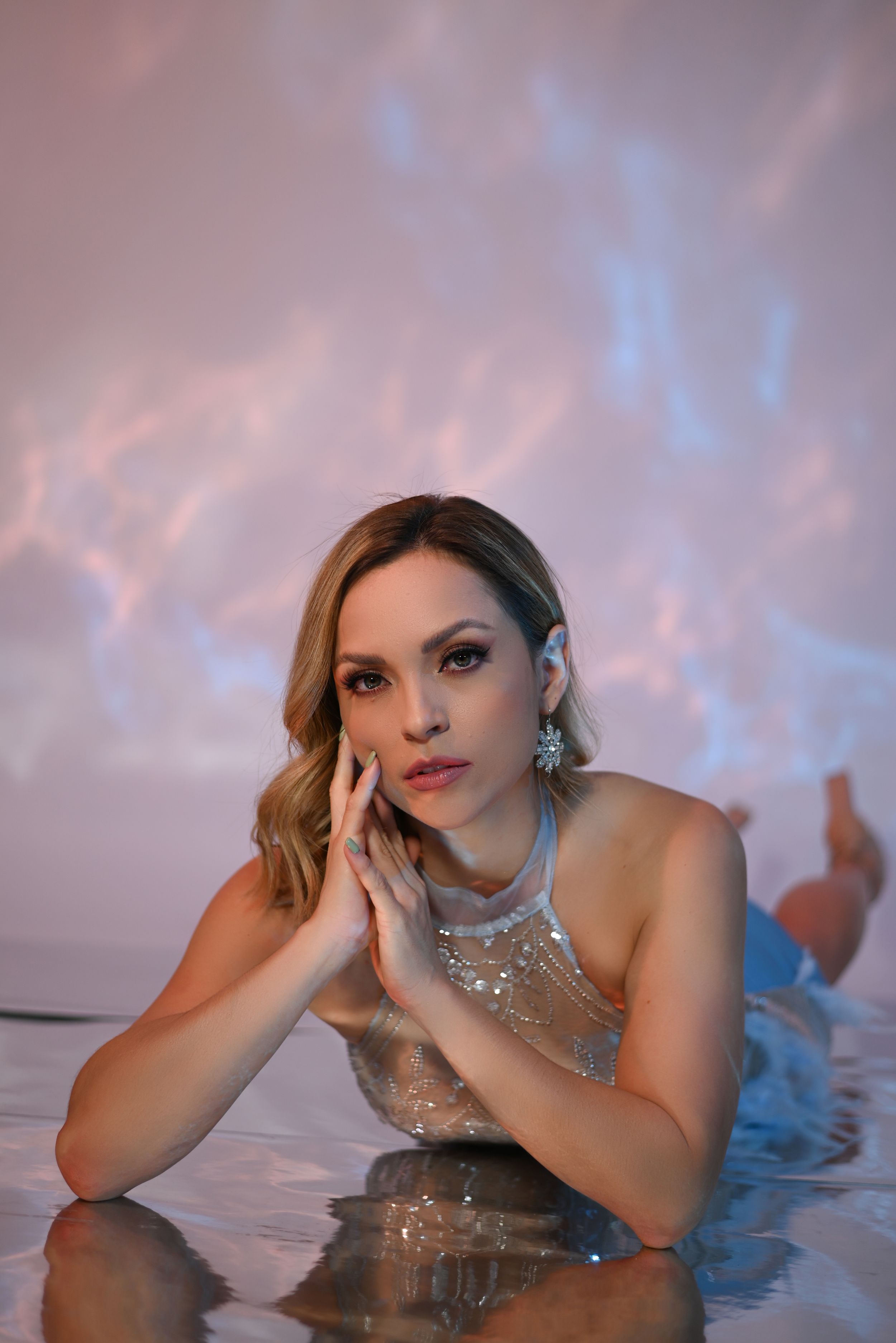 A woman in a sparkly dress poses against reflective water with a pink and blue sunset sky in the background.