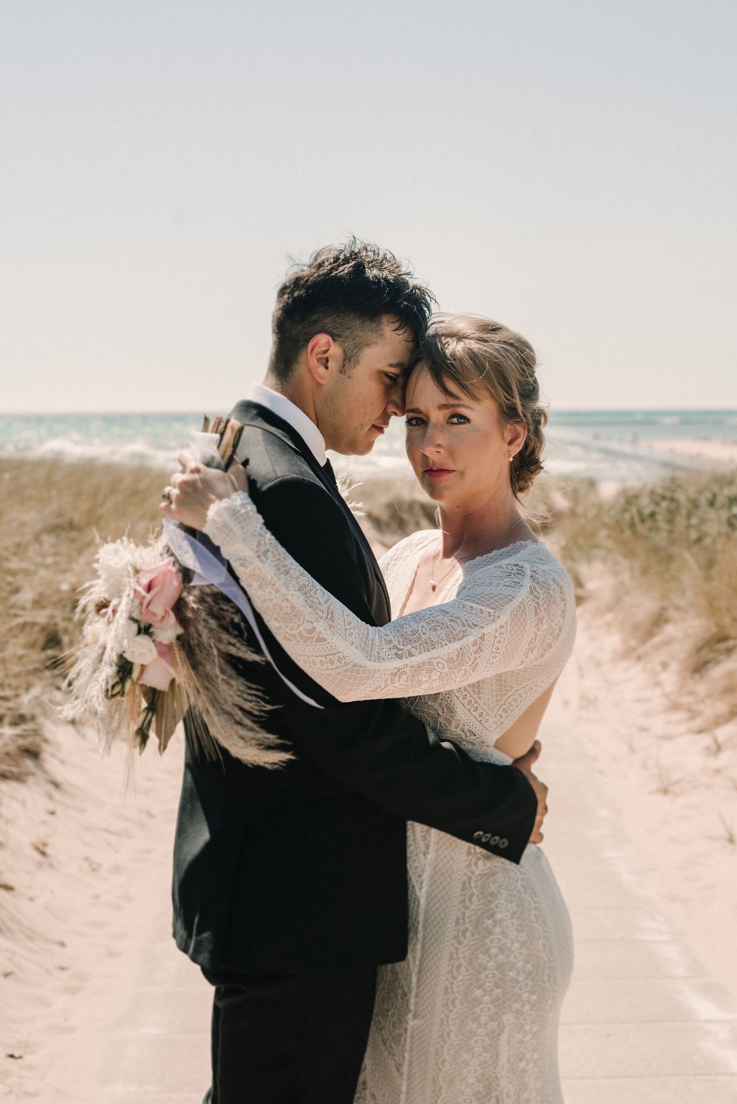A couple embraces on a sandy beach with dried pampas grass bouquet.