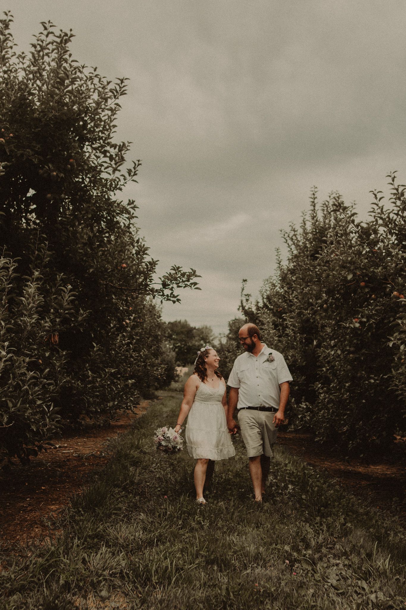 Romantic couple strolls through orchard in casual wedding attire.