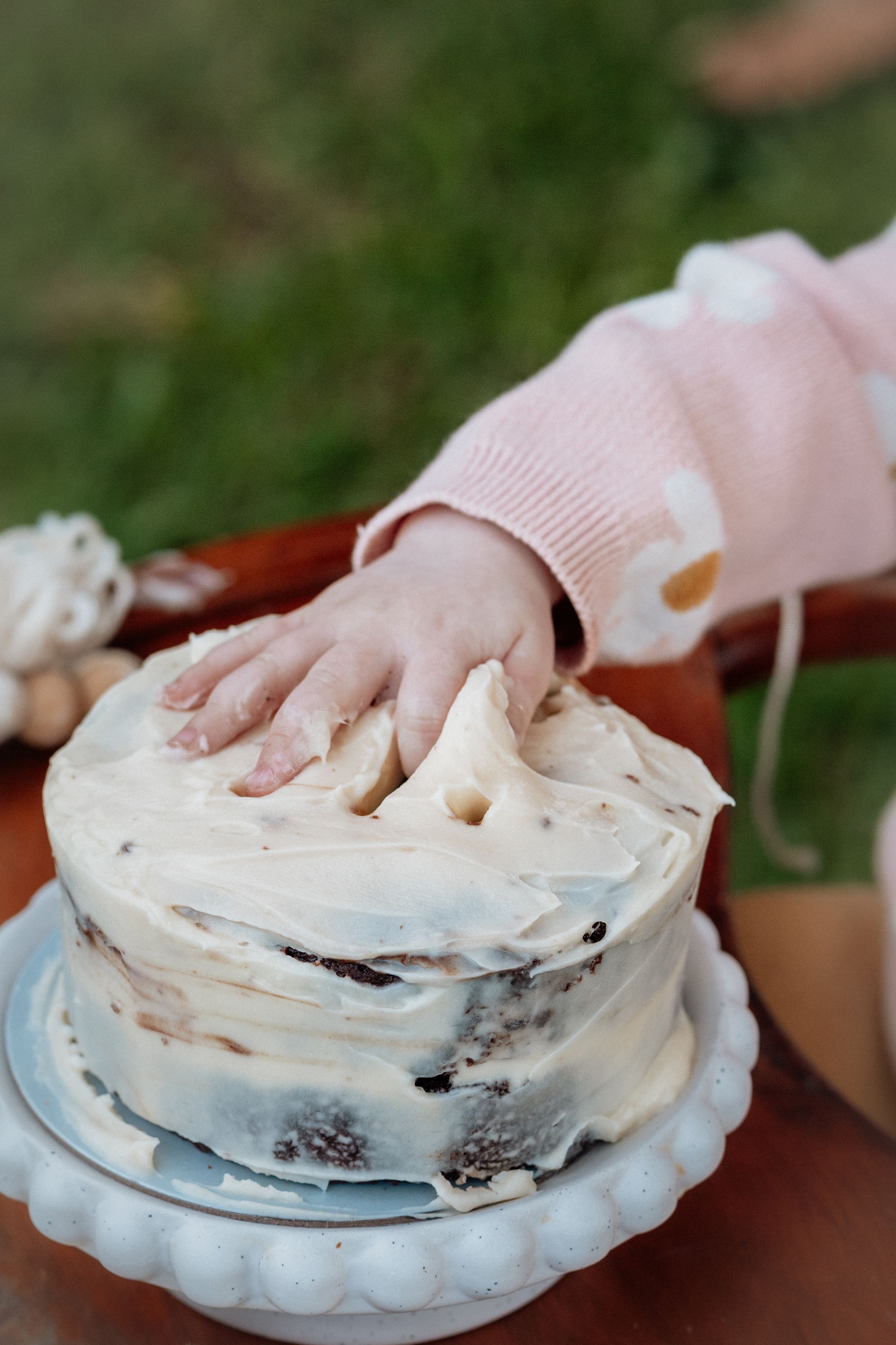 A baby's hand smashing into a white frosted smash cake on a cake stand during a first birthday celebration.