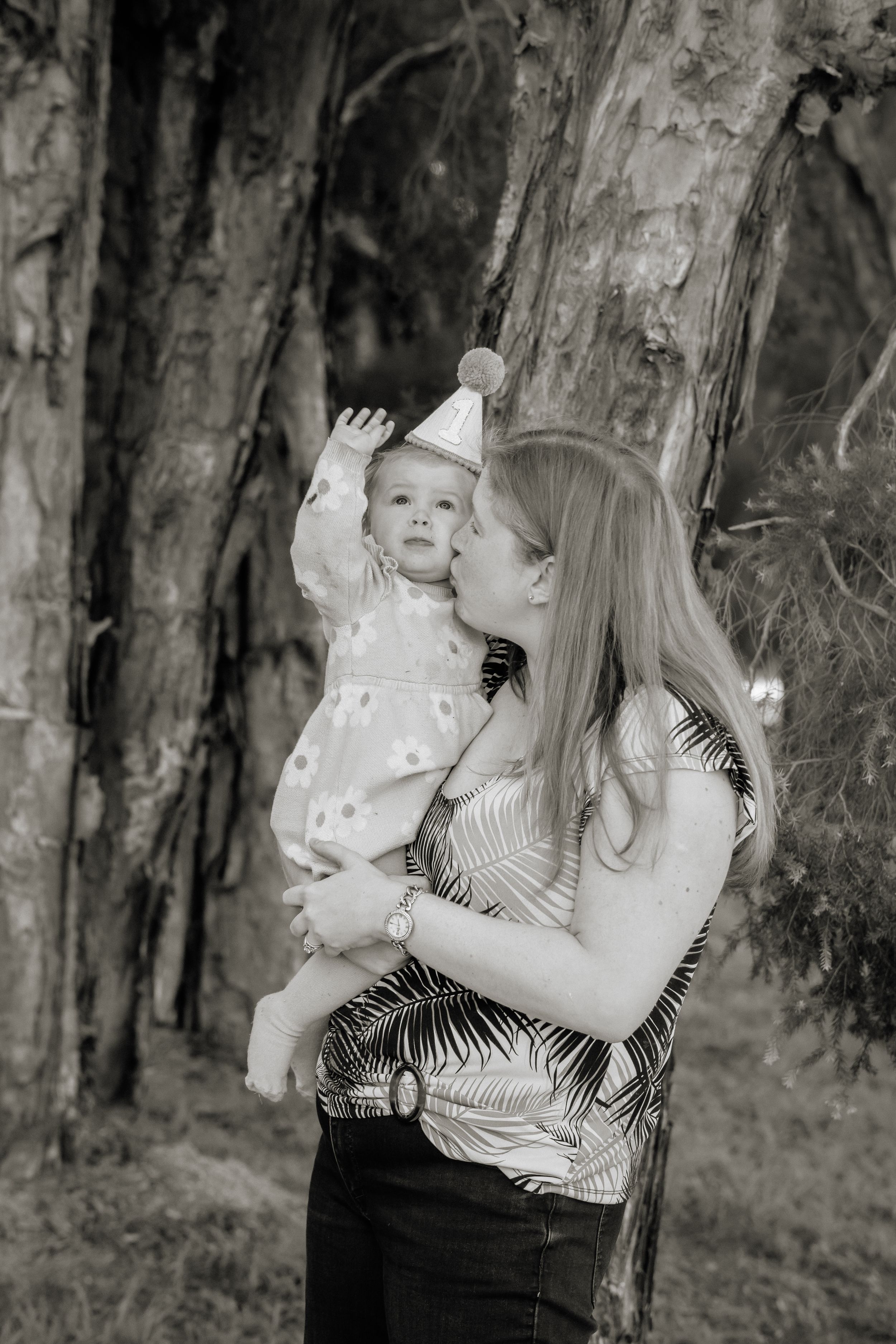 Black and white photo of a mother kissing her baby wearing a party hat outdoors near trees.