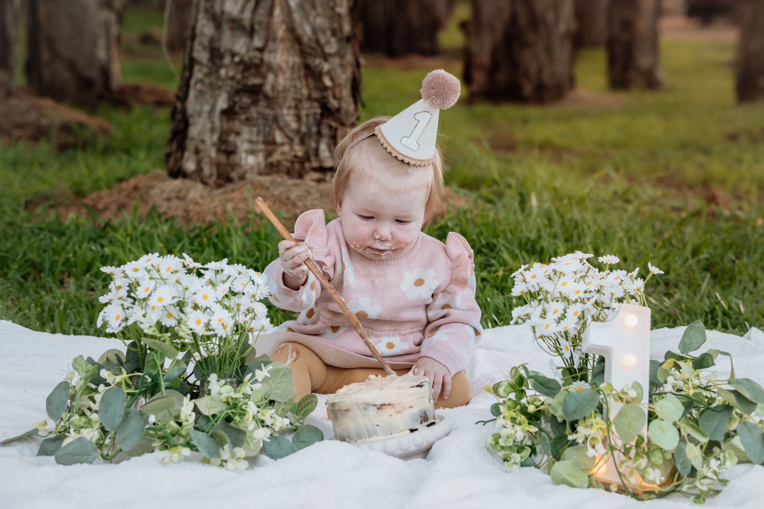 Baby girl in party hat celebrates first birthday on white blanket surrounded by white flowers and candles outdoors.