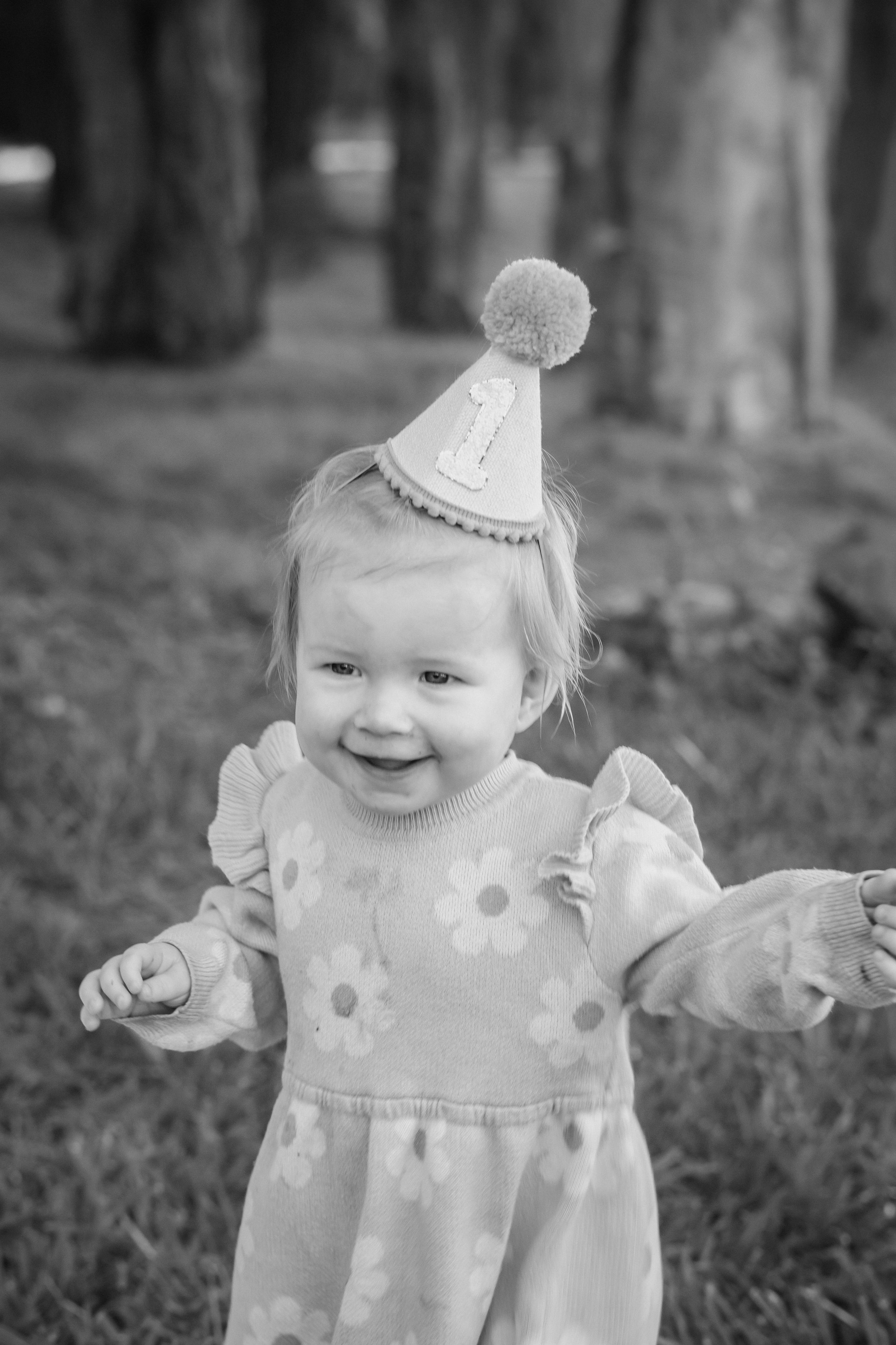 Black and white photo of a smiling toddler girl wearing a number 1 birthday hat and floral dress outdoors.