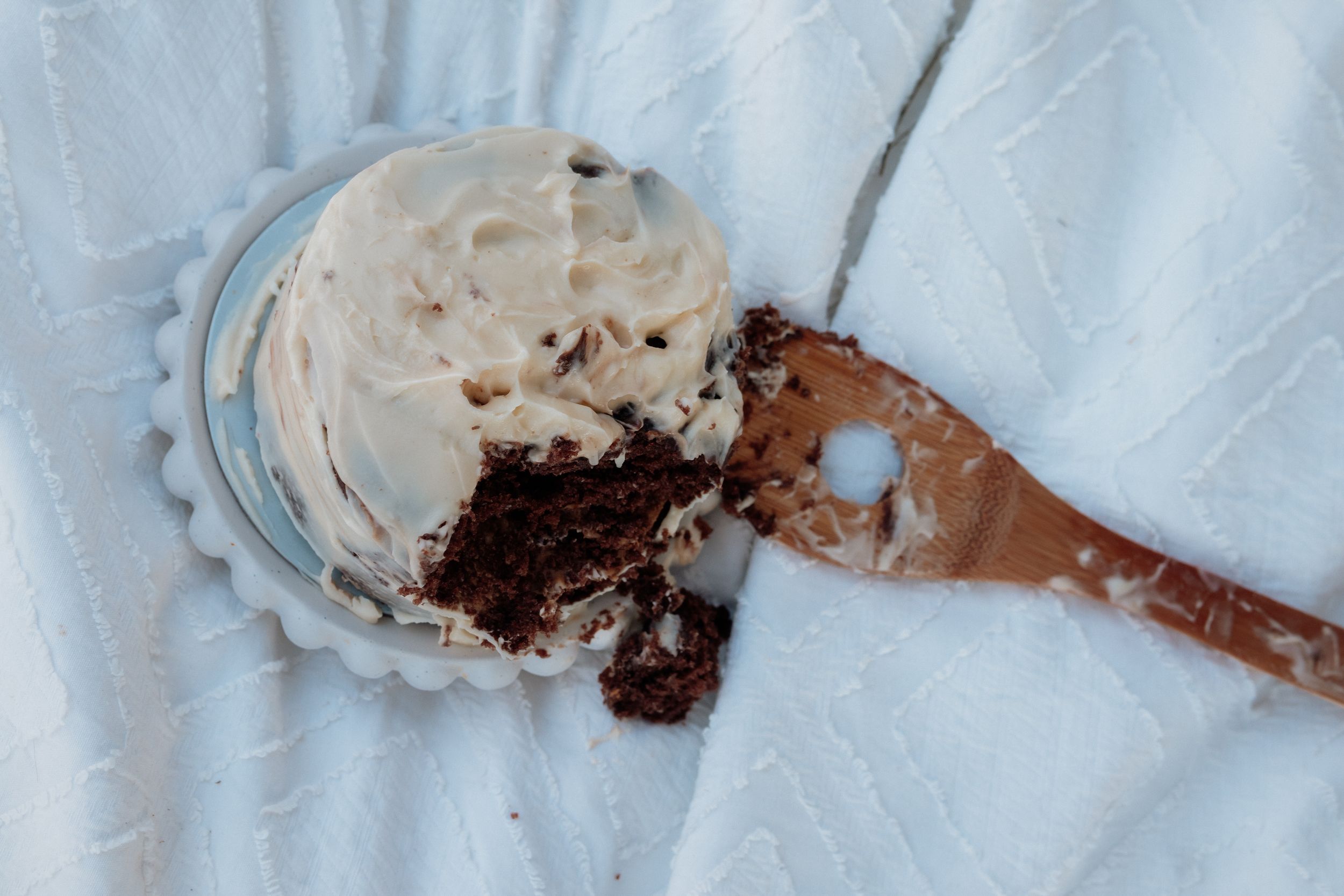 A partially eaten chocolate cupcake with white frosting on a decorative plate with a wooden spoon.
