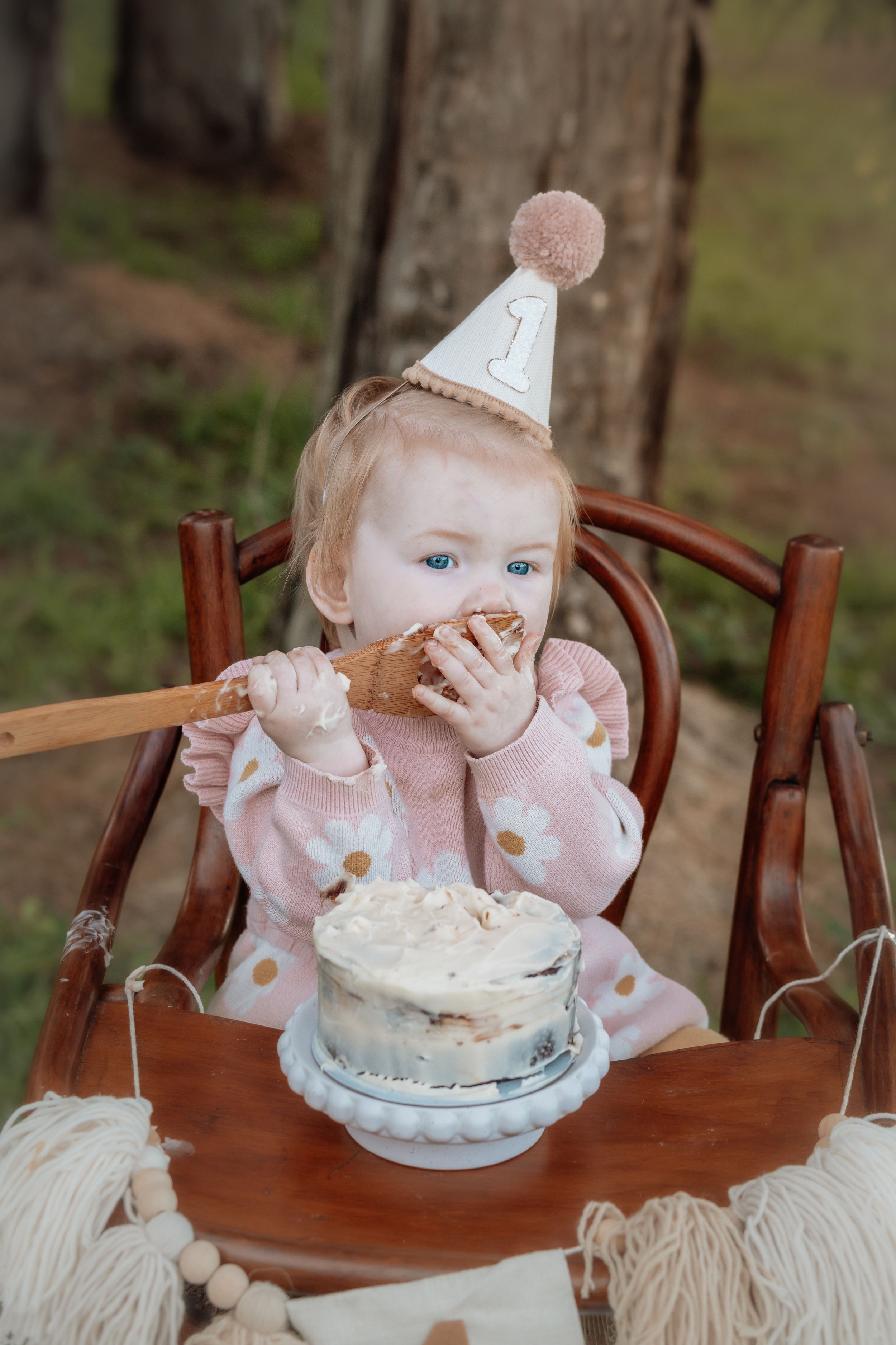 Baby girl in pink dress and number 1 party hat eating smash cake in wooden high chair outdoors.