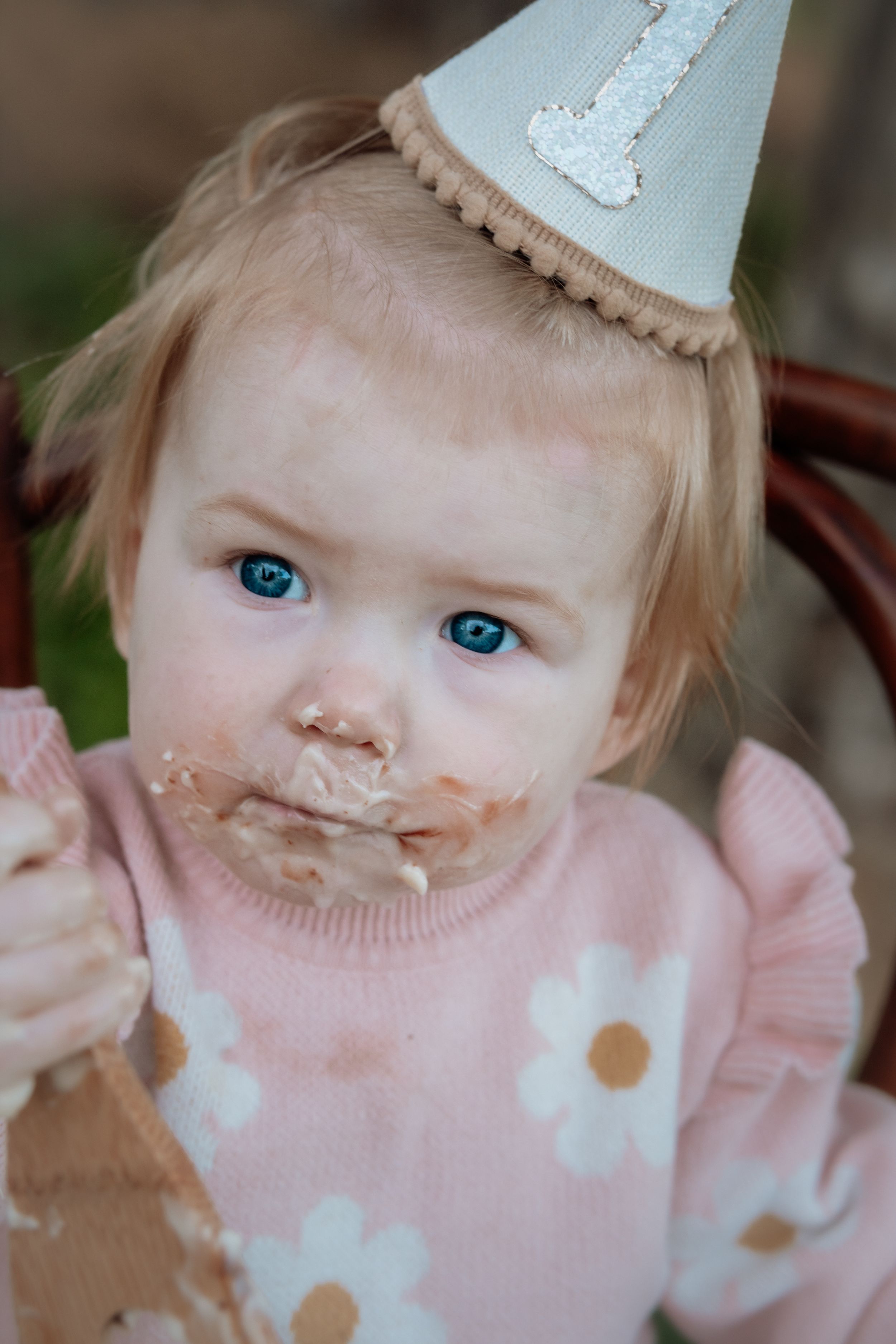 Messy baby girl with blue eyes celebrating her 1st birthday, wearing a party hat and pink floral sweater.
