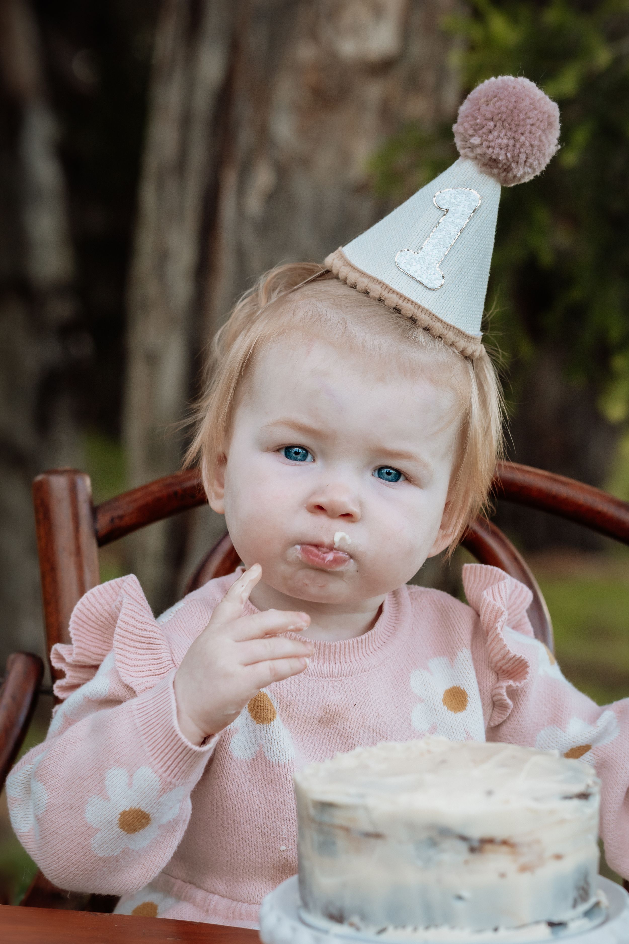 A baby girl in a pink floral sweater and birthday hat enjoys her first birthday cake outdoors.