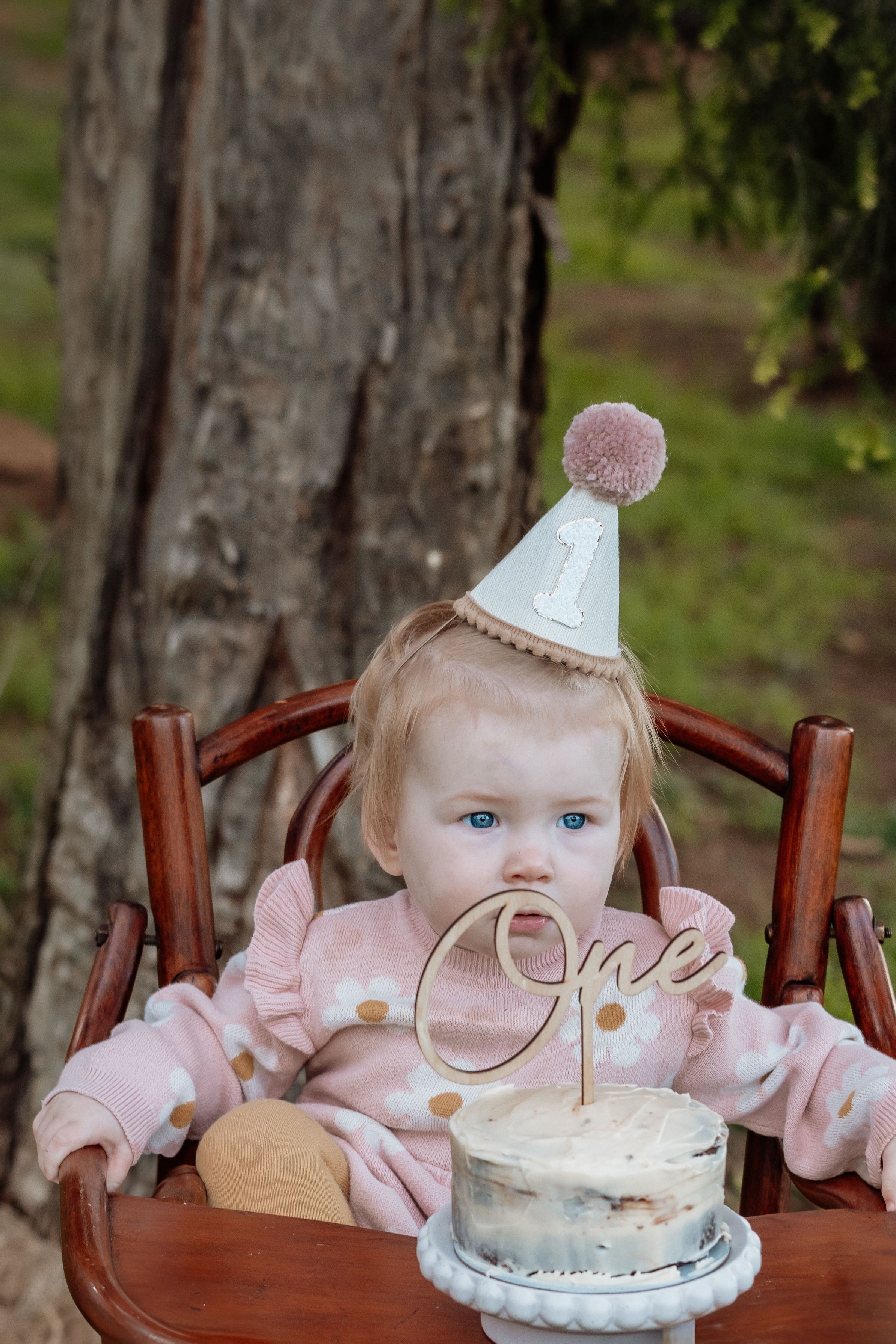 Baby girl in pink outfit and number 1 party hat sitting with first birthday smash cake outdoors.