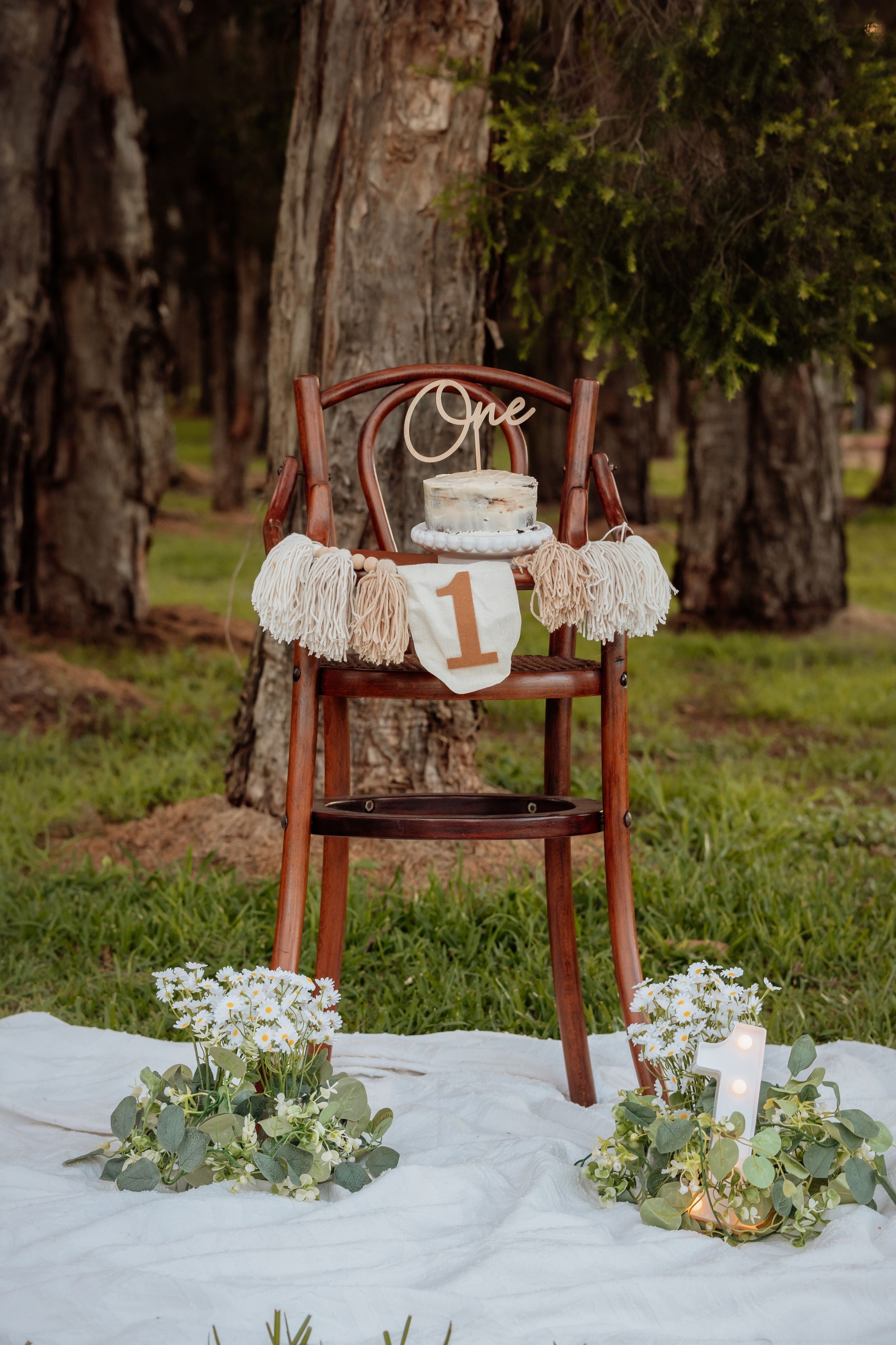 Rustic first birthday setup with a wooden high chair, smash cake, macrame garland, and floral arrangements on a white blanket outdoors.