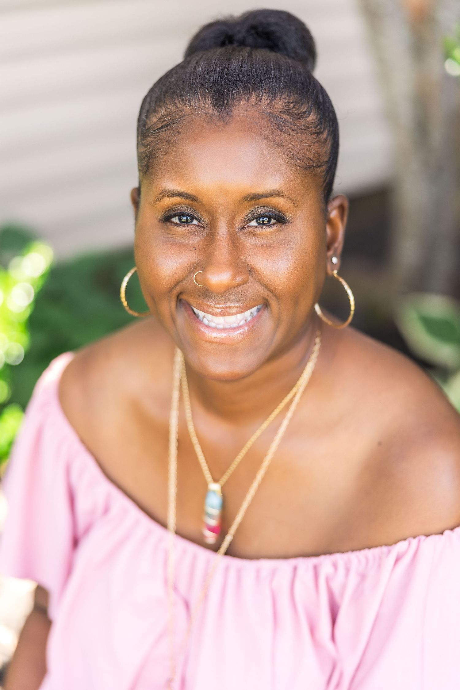 Smiling woman wearing a pink off-shoulder top with gold jewelry against a blurred outdoor background.