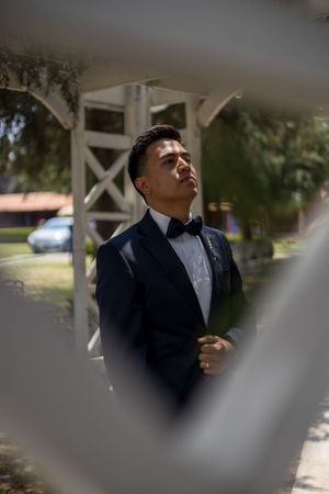 A groom in a classic black tuxedo and bowtie poses outdoors in natural lighting.
