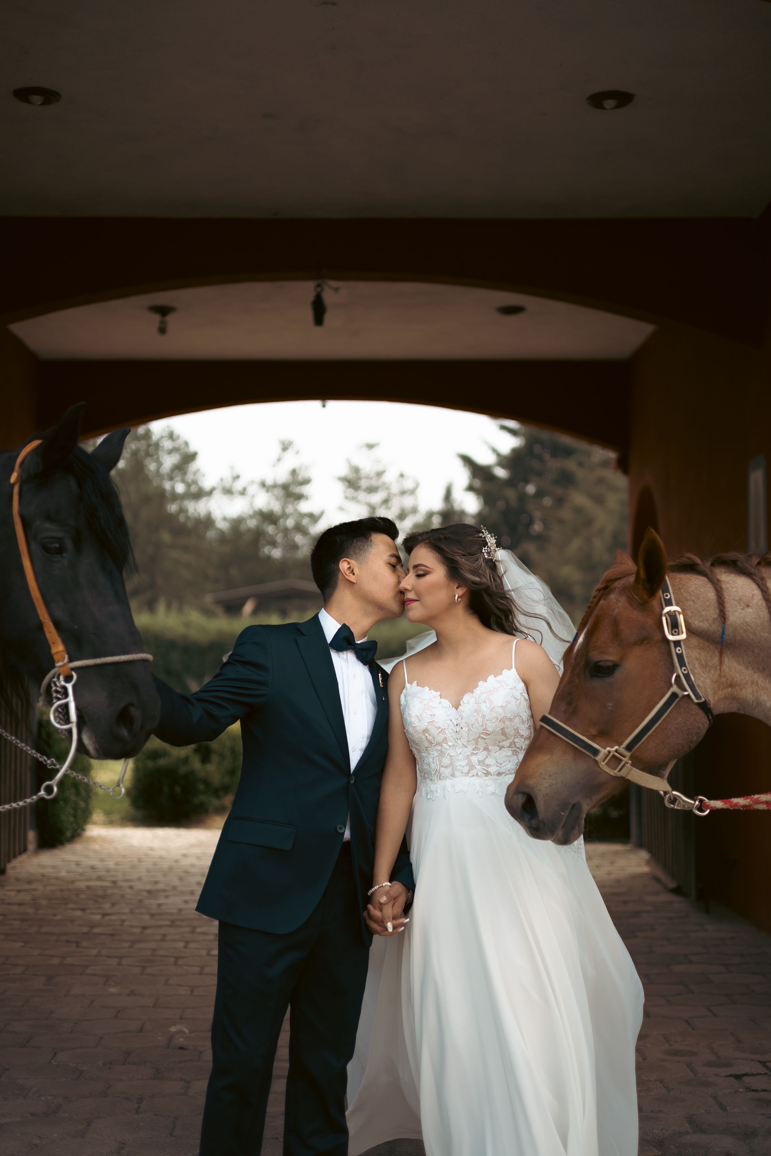 Wedding couple posing with horses in a rustic stable setting.
