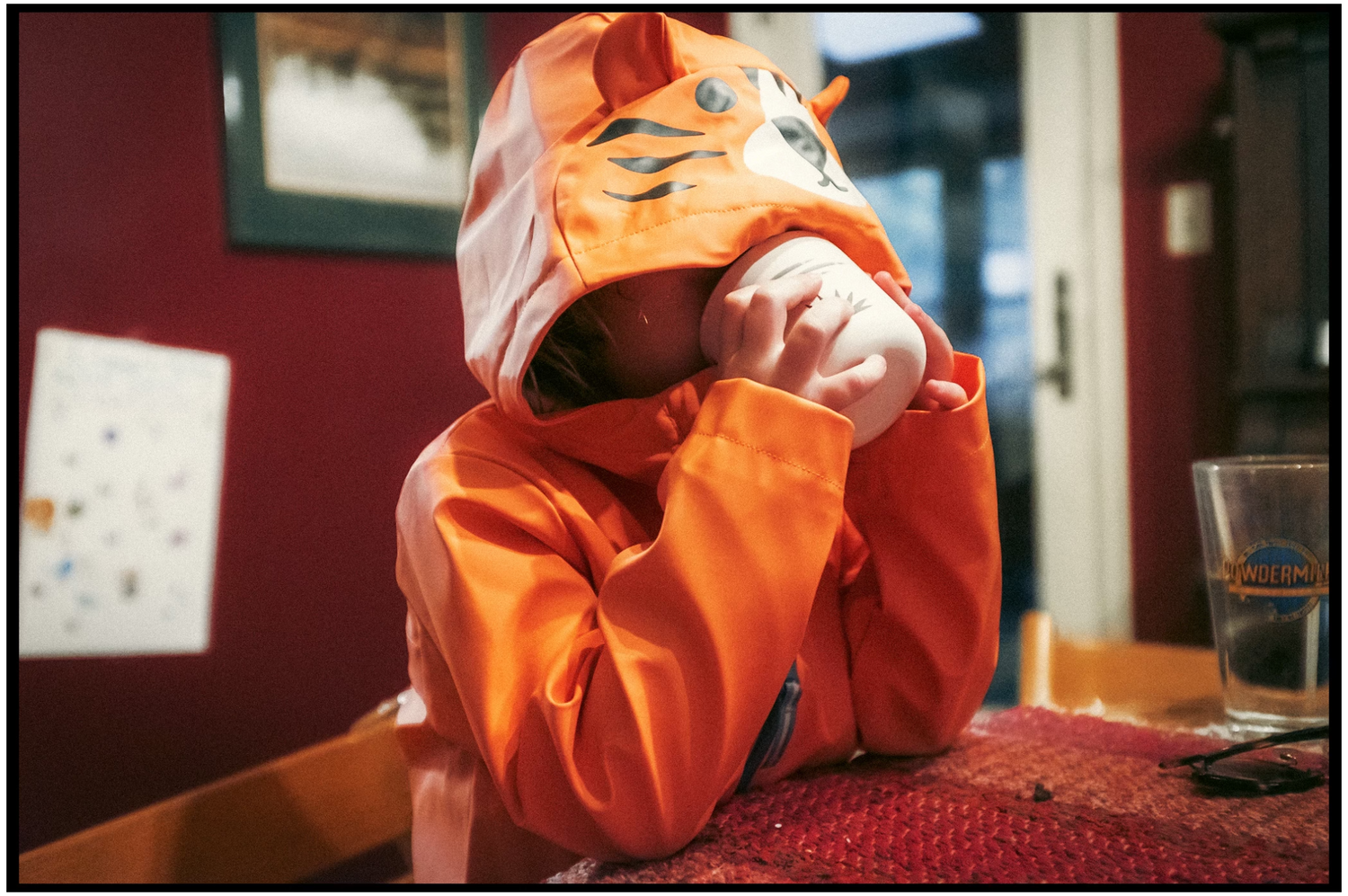 Person wearing a bright orange tiger hooded garment leans on table in indoor setting.