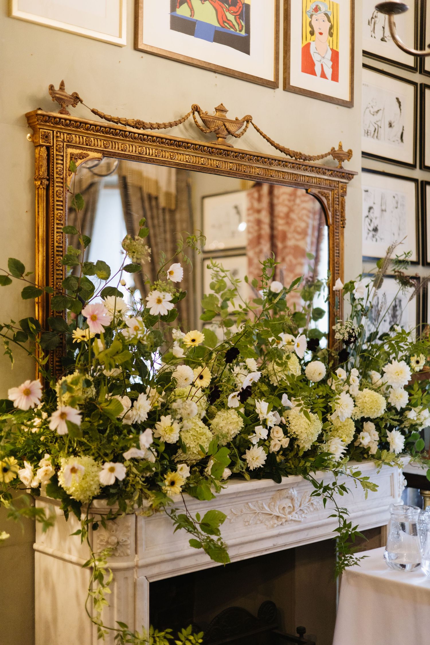 Elegant floral arrangement with white blooms sits atop fireplace mantel beneath ornate gold mirror.