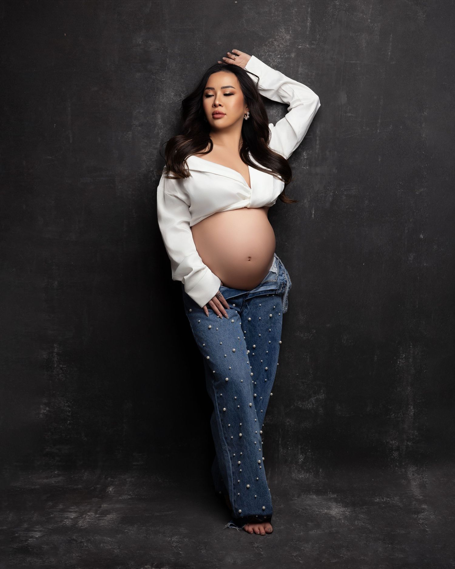 Maternity portrait of expectant mother in white button-down shirt and pearl-studded jeans against dark backdrop.